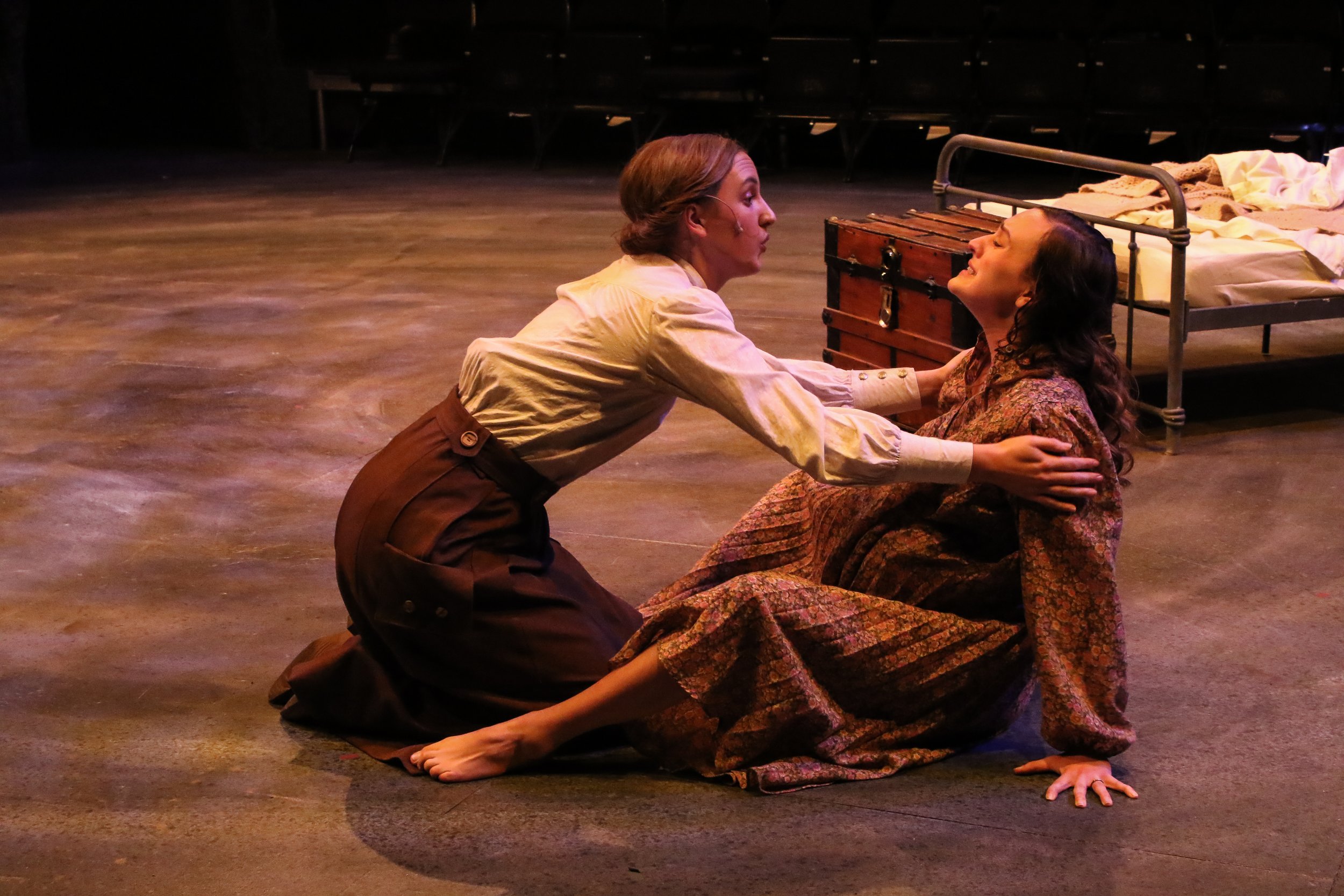 Two women are on stage, engaged in an emotional performance. One woman is kneeling, holding the other woman who is sitting and leaning back on her hands, both making intense facial expressions. The background is dark with a bed and a chest nearby.