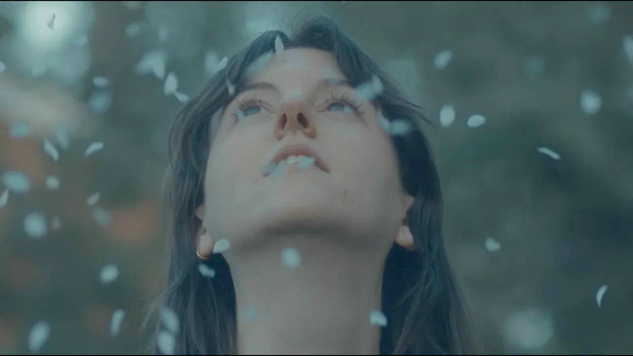 A close-up of a woman with dark wet hair, looking upward with a serene expression, surrounded by falling white flower petals or confetti.