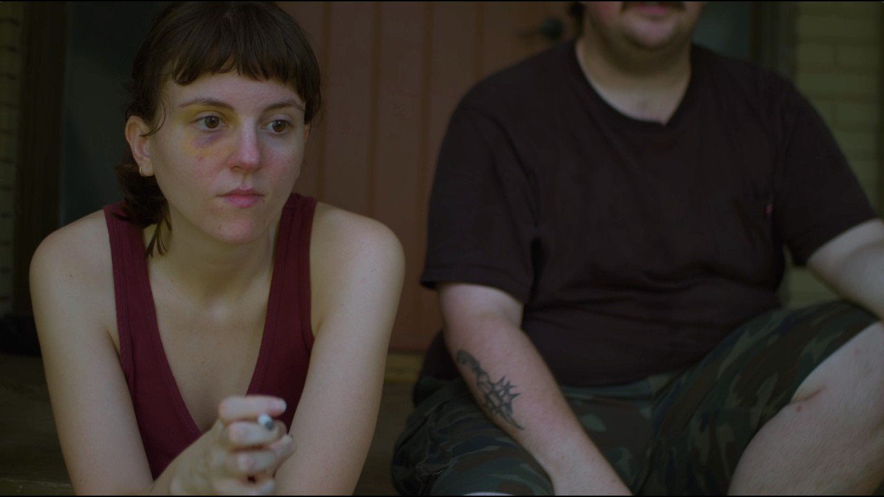 A young woman with short brown hair, wearing a red tank top, looking contemplative with a bruise under her eye, sitting next to a man with a tattoo on his forearm, wearing a black t-shirt and camouflage shorts.