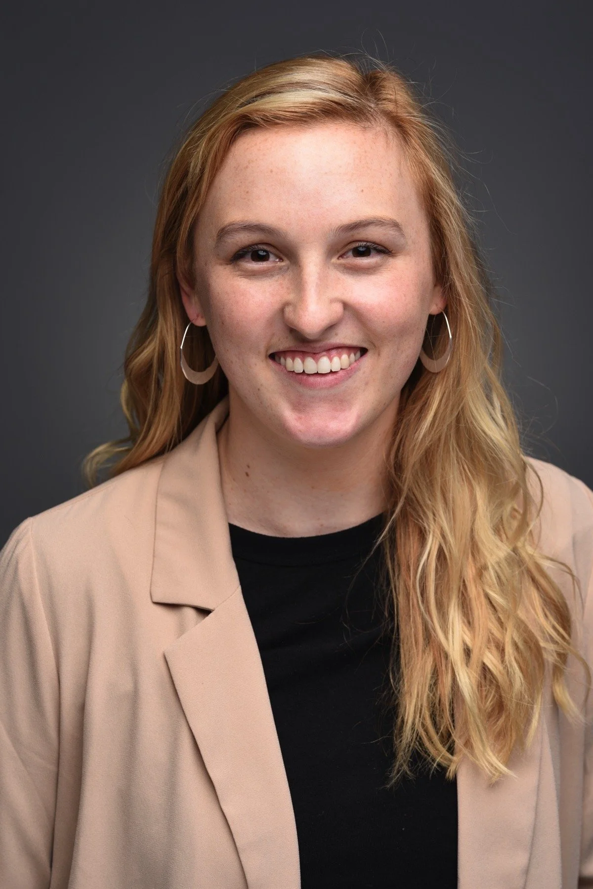 A woman with long, wavy blonde hair and hoop earrings, smiling at the camera, wearing a black top and a beige blazer, against a dark gray background.