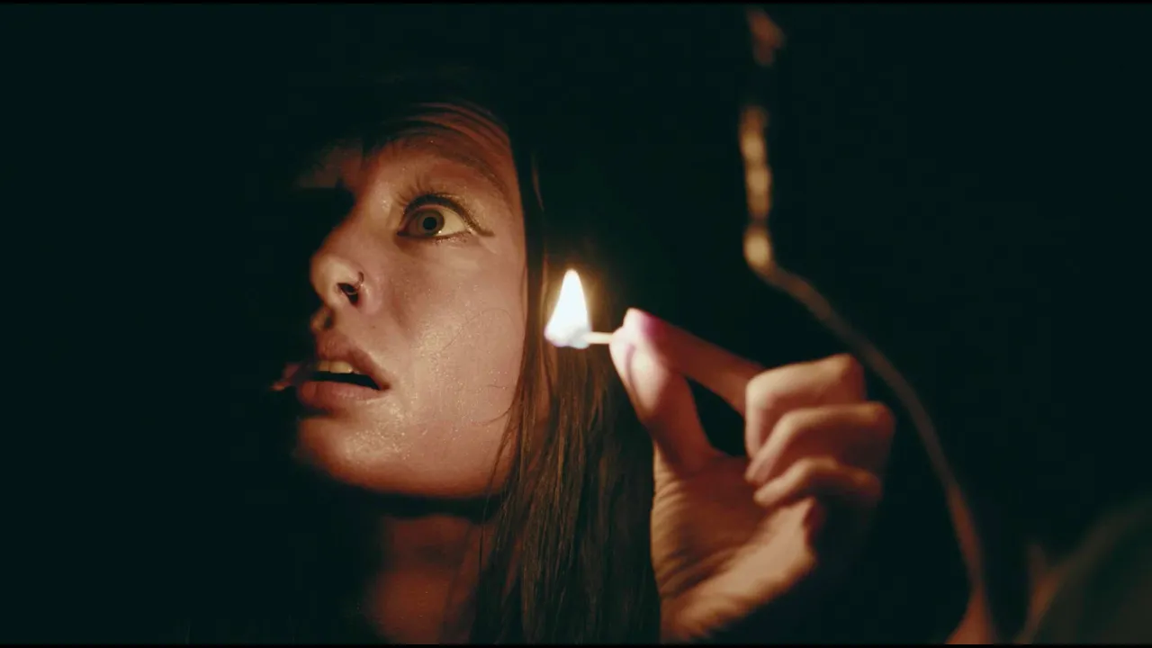 Close-up of a person with pale skin and long dark hair holding a lit match near their face, in a dark environment.