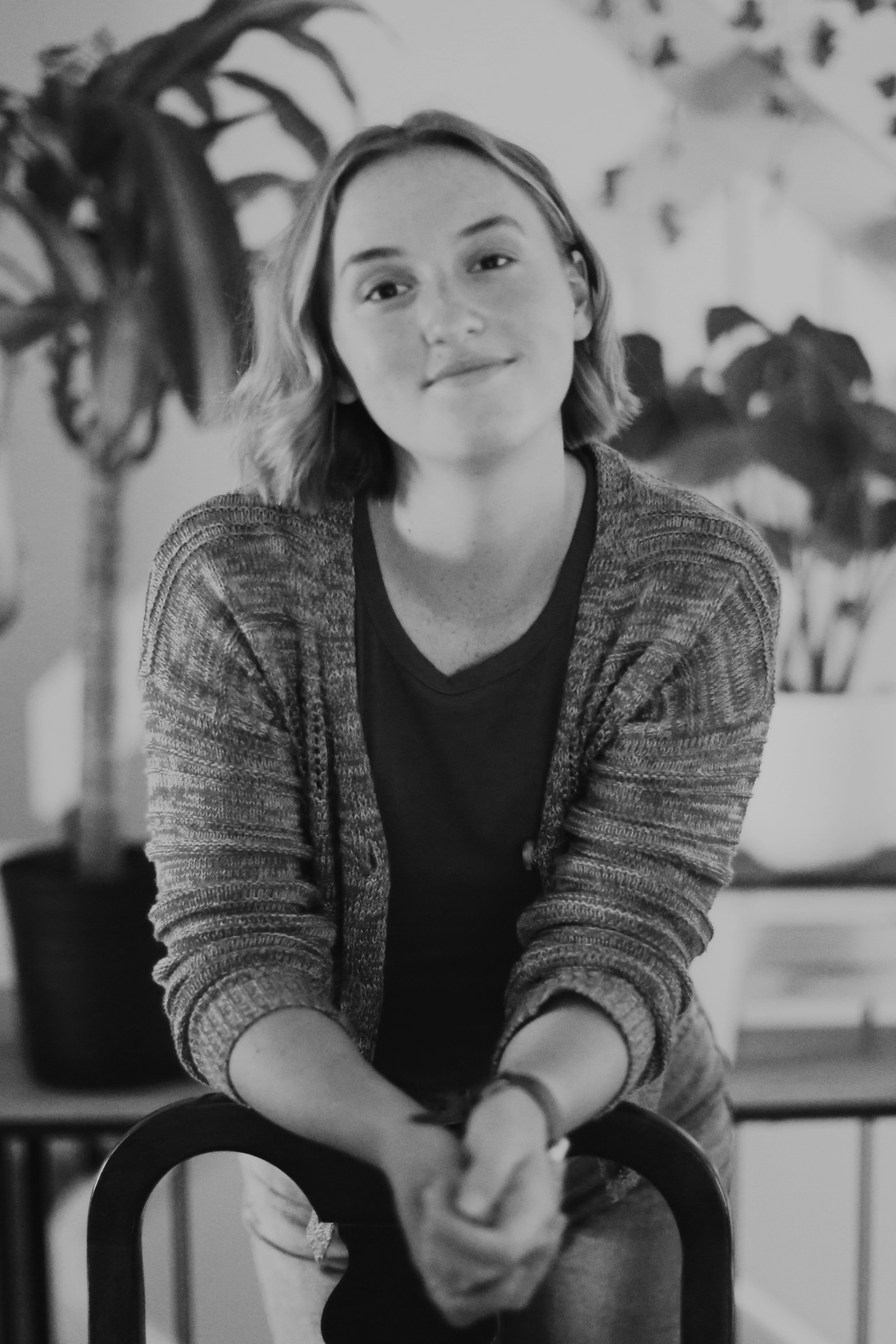 A headshot of Laura Falcione: a woman with shoulder-length hair is smiling and sitting at a table, holding her hands in front of her, with plants in the background.