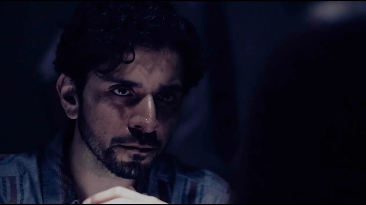A man with dark, curly hair and a beard sitting in a dark environment, looking intently at something off screen.