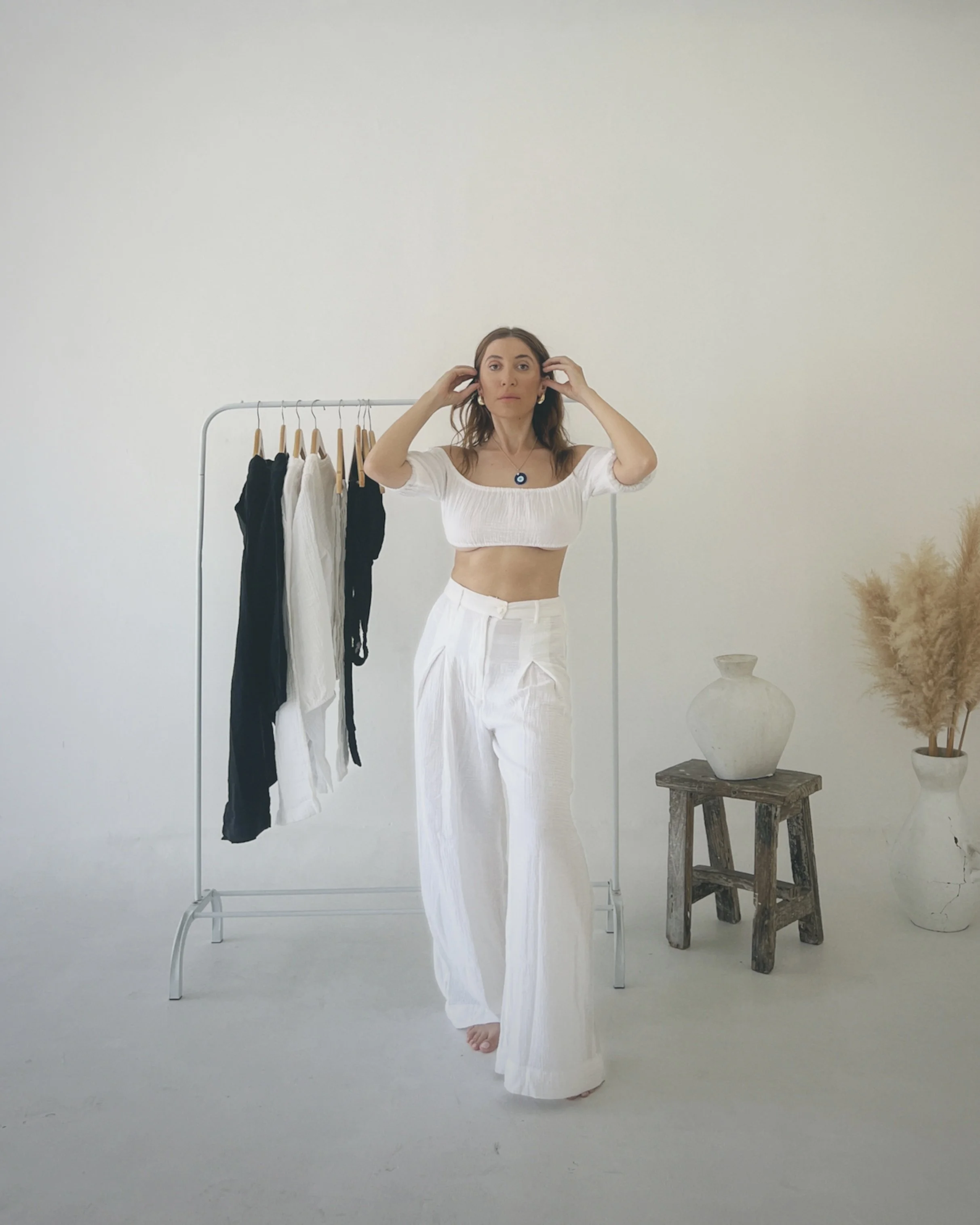 A woman in white pants and a white crop top standing next to a clothing rack with black and white clothing, in a minimalistic room with a vase and dried pampas grass on a rustic wooden stool.