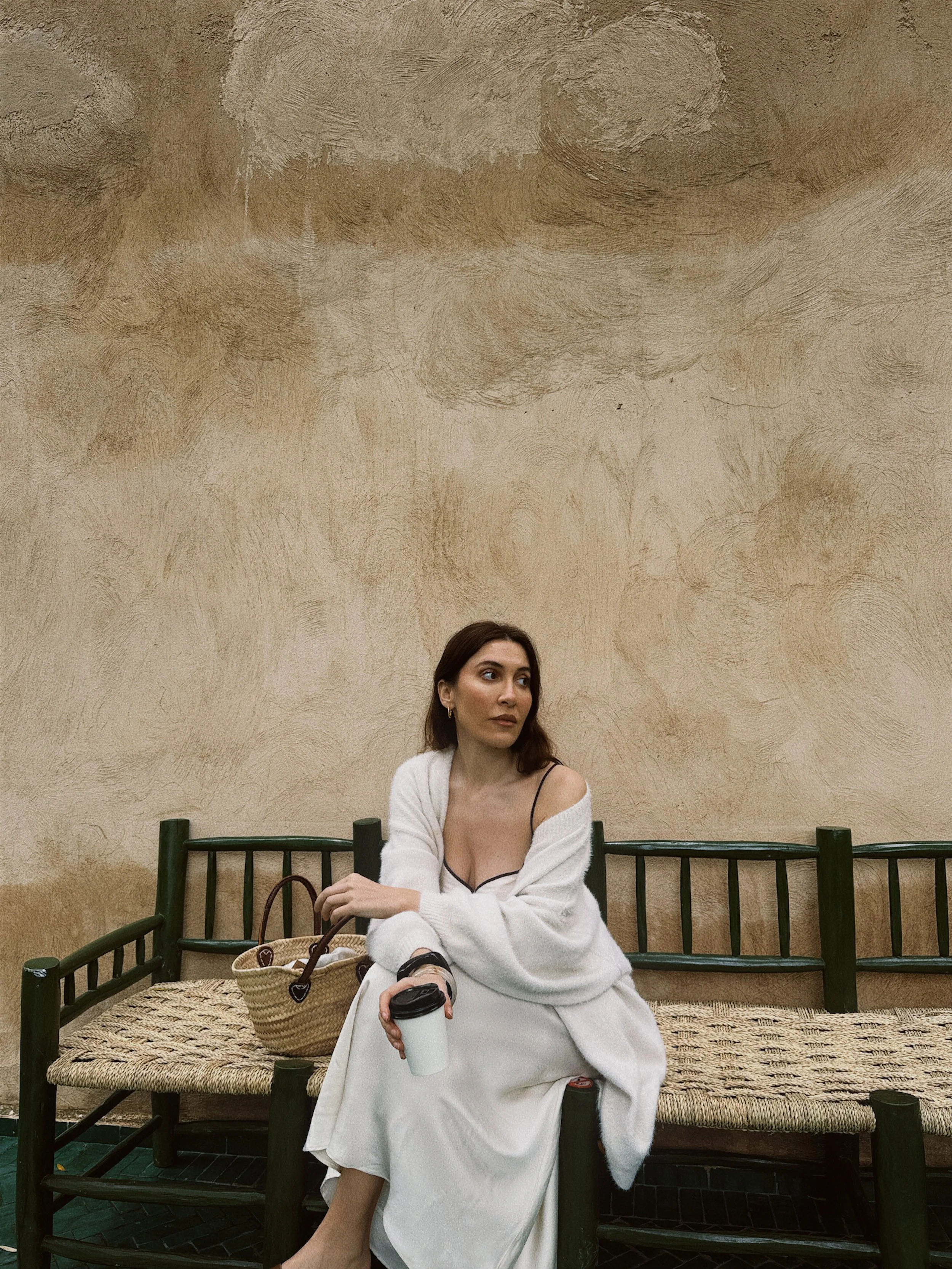 A woman with long brown hair, wearing a black camisole and a white cardigan, sitting on a dark green wooden bench with a woven seat. She is holding a disposable coffee cup in her right hand and has a straw bag next to her. She appears to be looking to the side in a contemplative pose, against a textured beige wall.