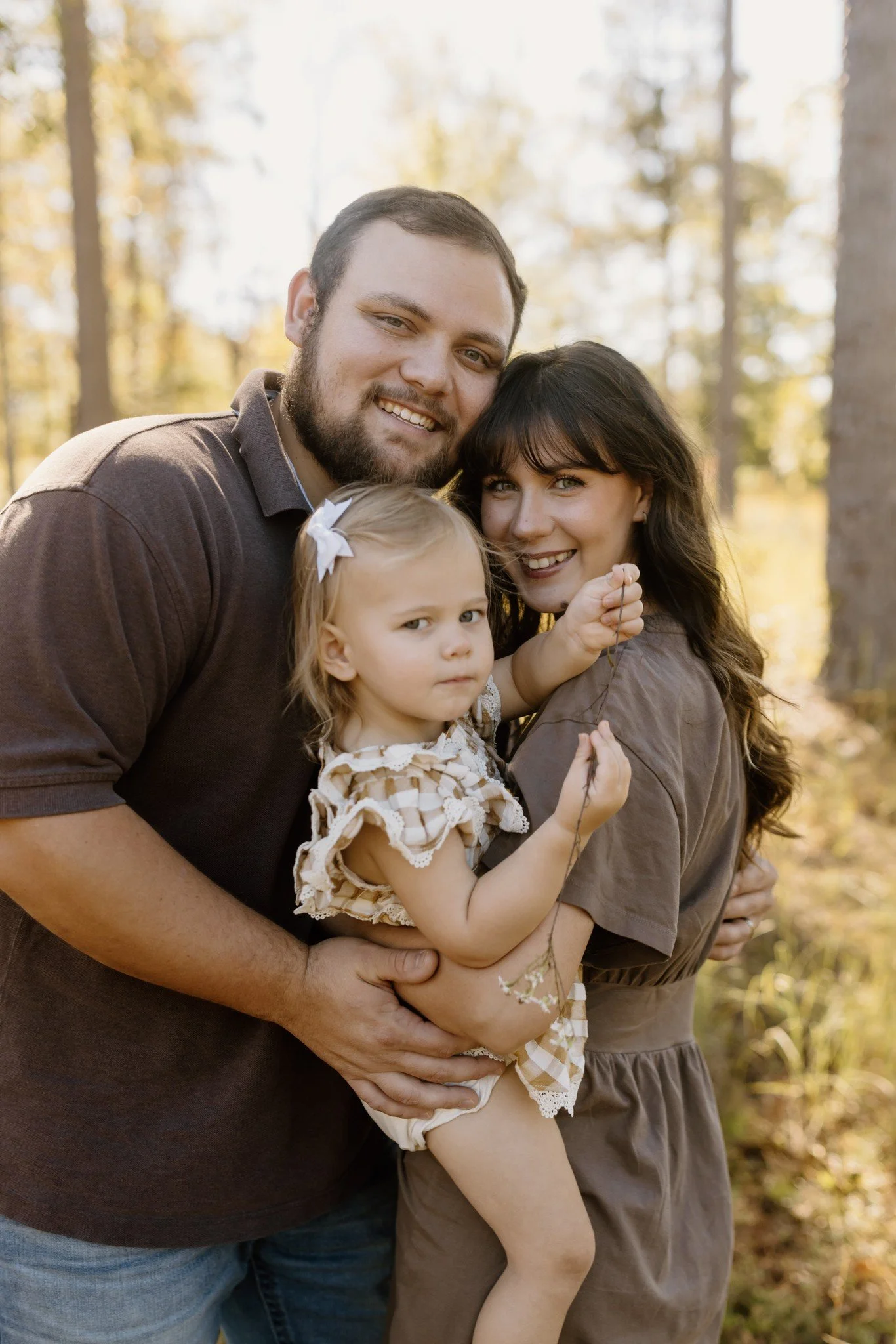 Mom, dad, and their little girl hug and smile at the camera in warm sunlight under tall pines