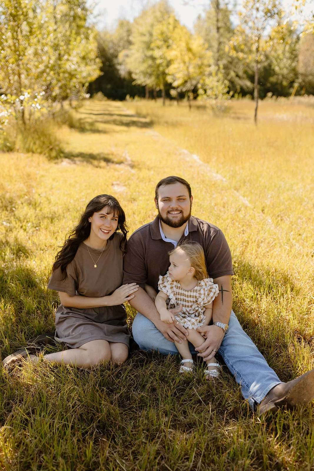 Smiling family sits together in tall golden grass with their little girl on dad's lap