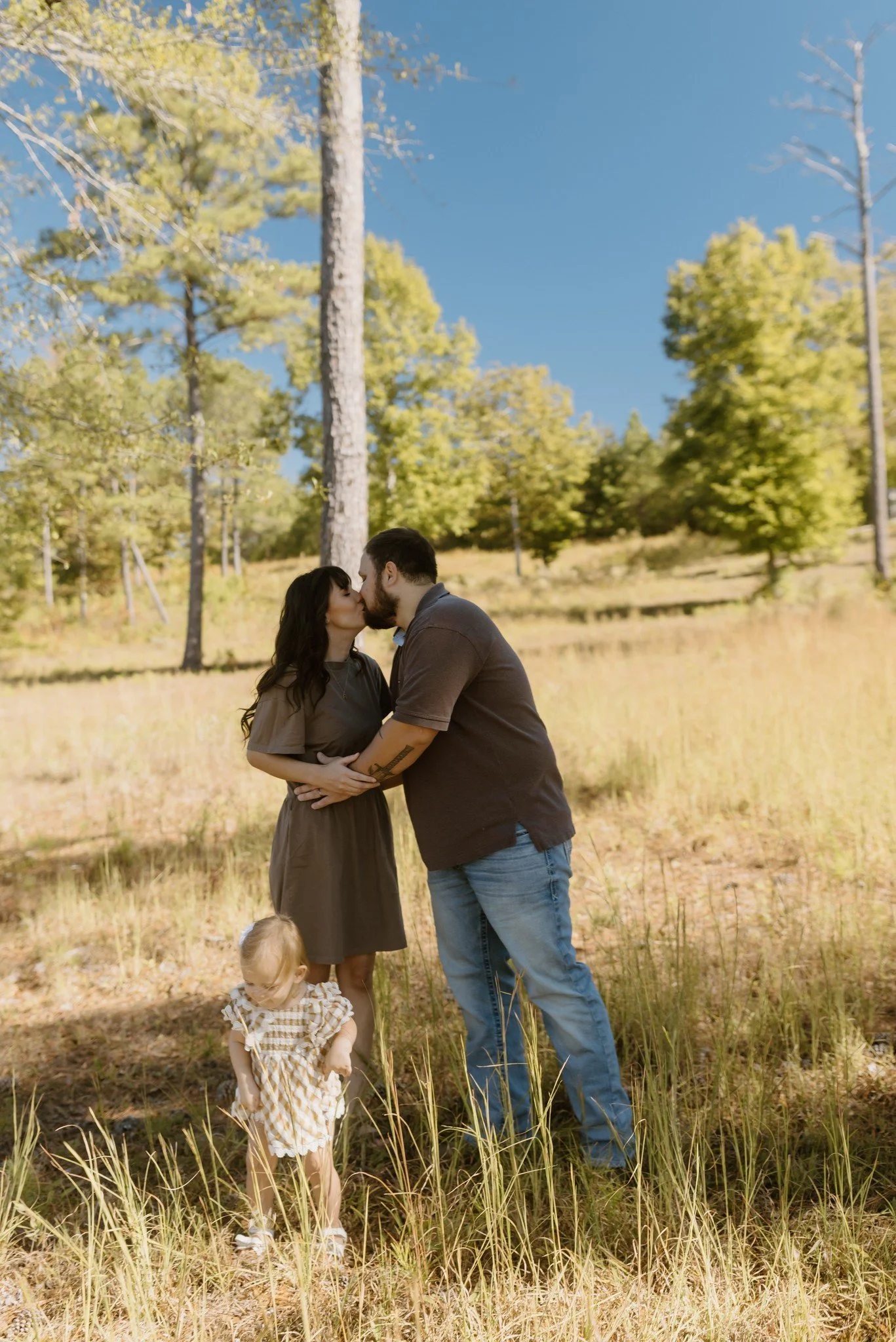 Parents share a kiss in a grassy fall field while their toddler in a gingham dress explores the grass