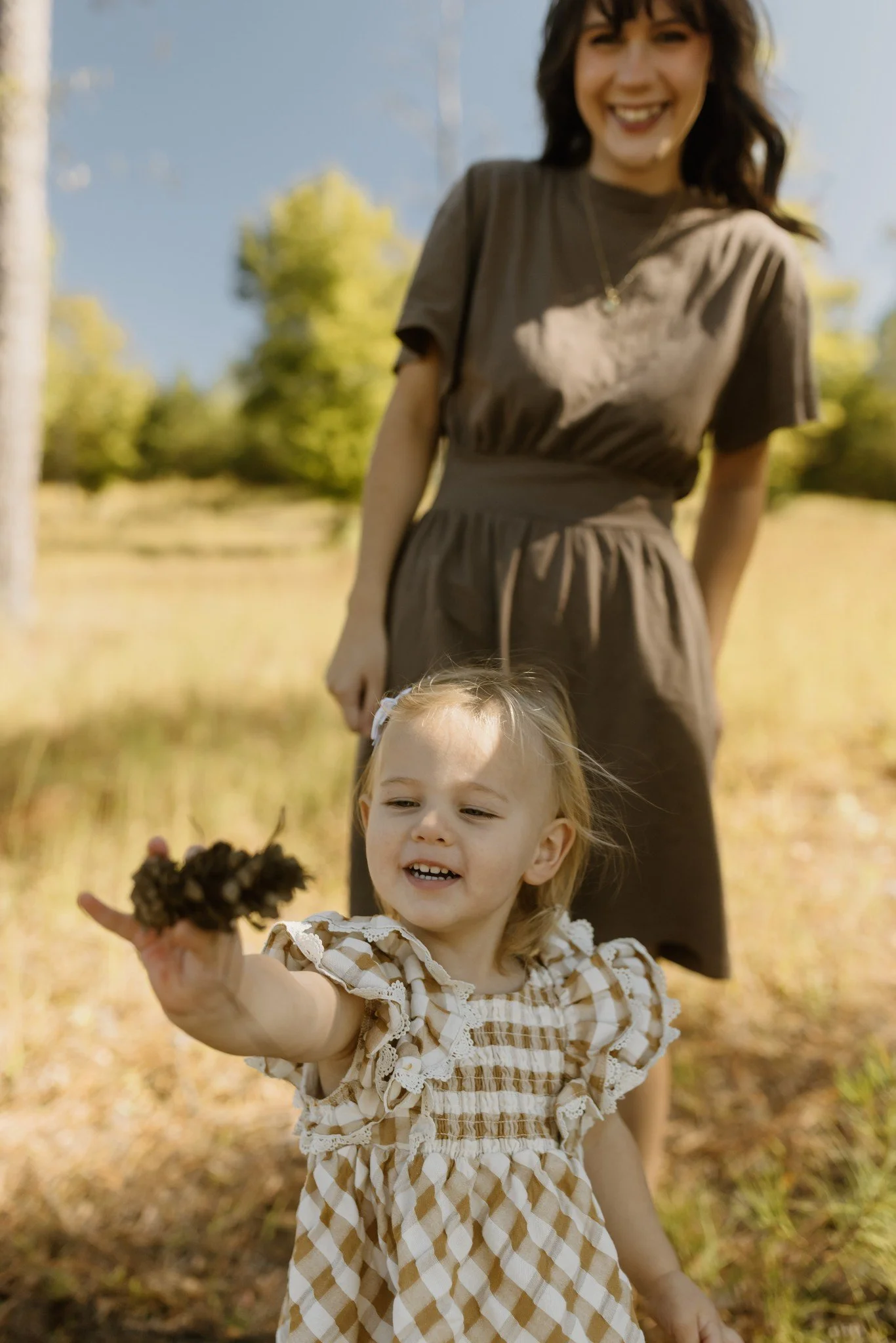 Little girl proudly holds a pinecone toward the camera while her mom smiles behind her