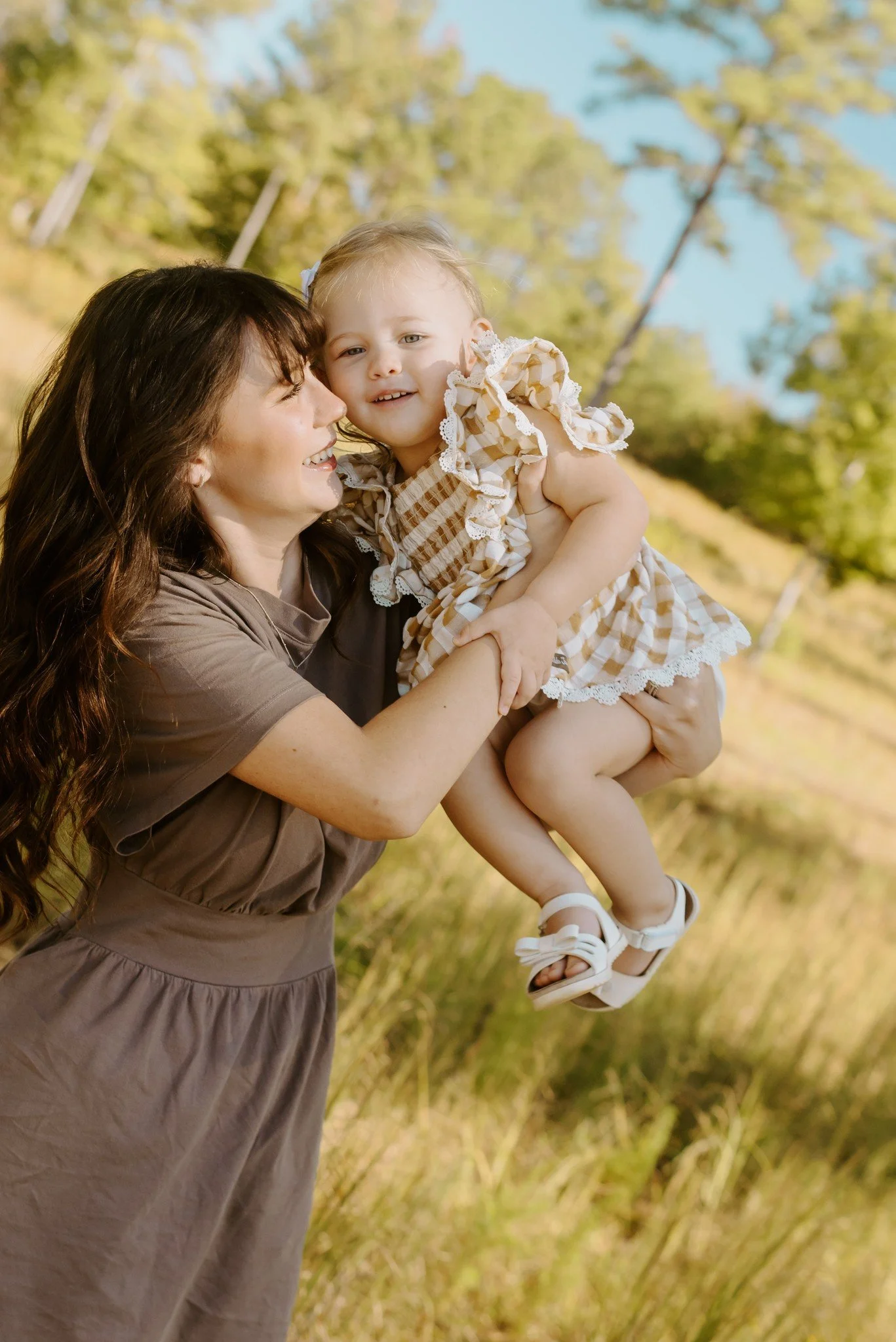 Mom swings her daughter up into the air as they both laugh on a grassy hill