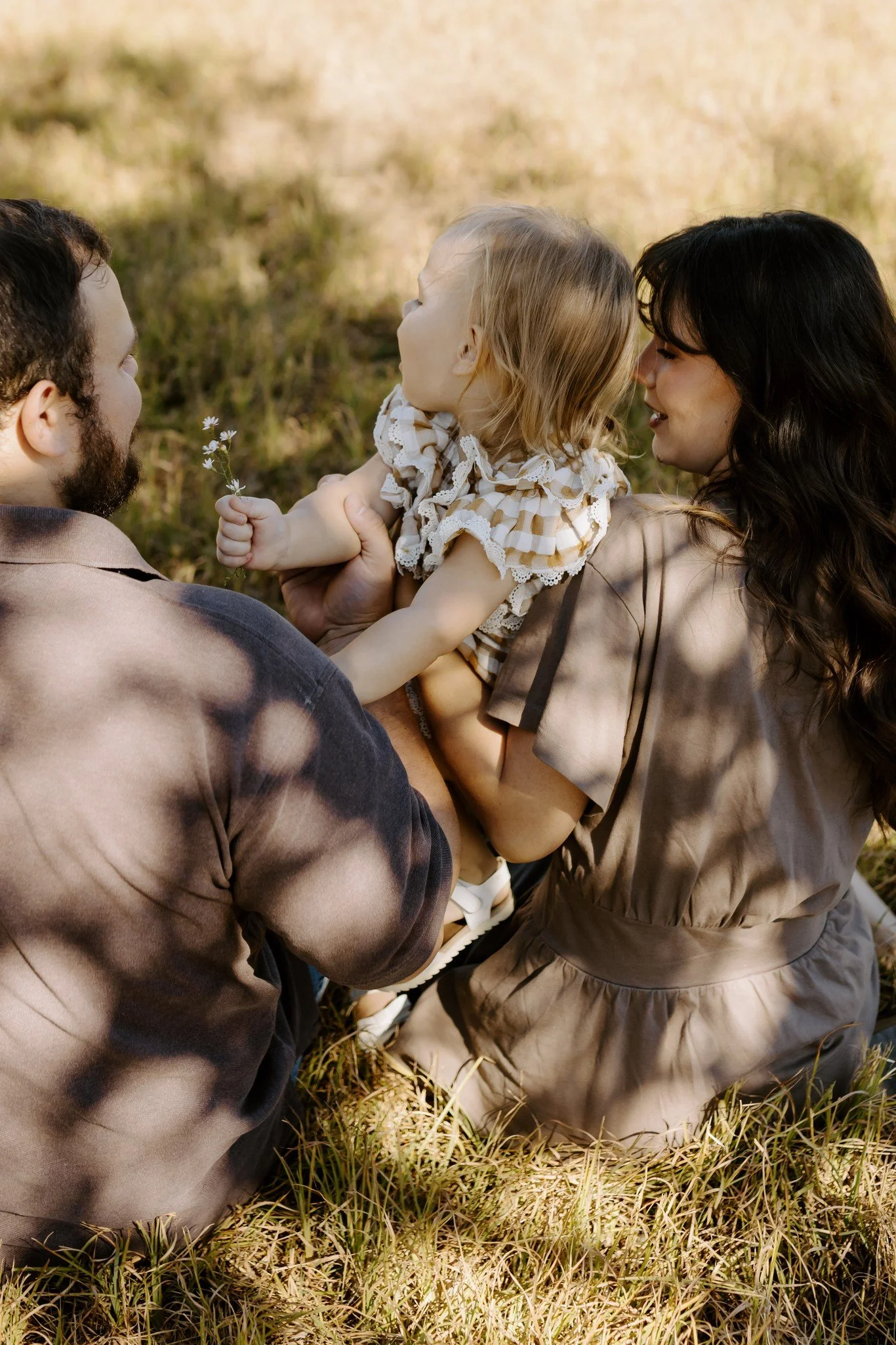 Seen from behind, a toddler holding a tiny wildflower turns toward her mom as the family sits in dappled shade