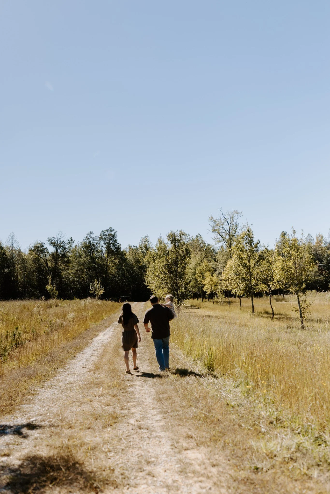 Parents walk down a sandy dirt road with their little girl, surrounded by golden fall grass and trees