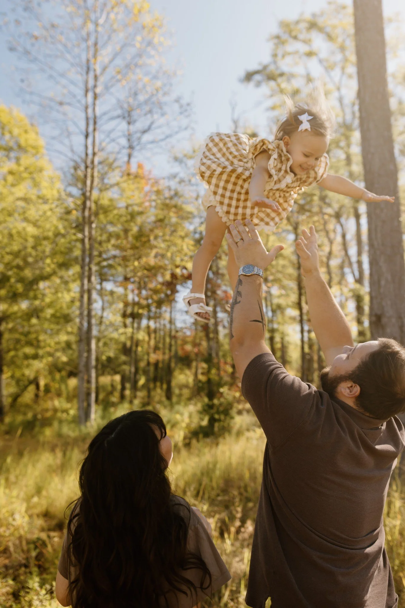 Dad tosses his giggling daughter into the air among tall pines while mom watches