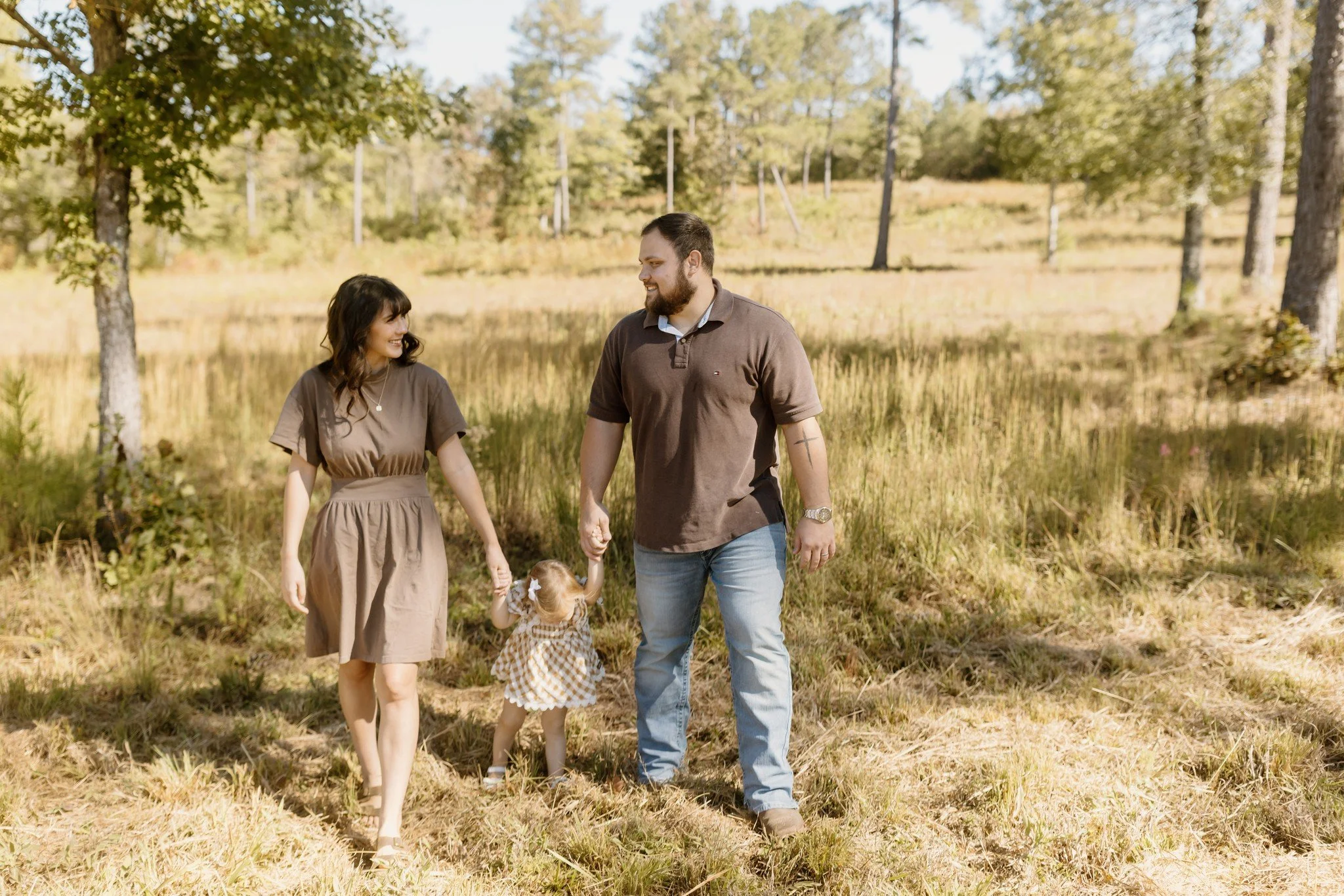 Mom, dad, and their toddler daughter walk hand in hand through a sunny field of tall fall grass