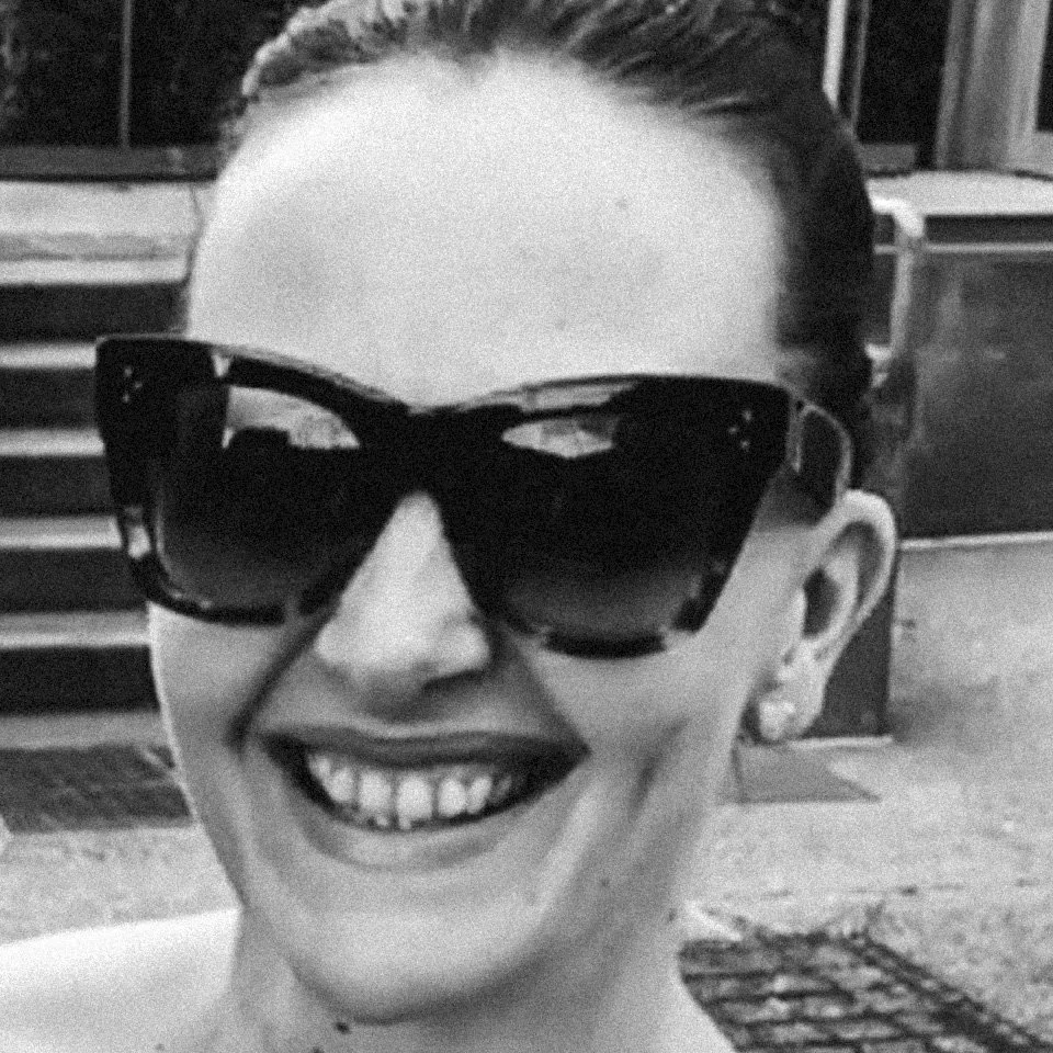 A woman smiling while wearing large dark sunglasses, with her hair pulled back. The photo appears to be black and white.