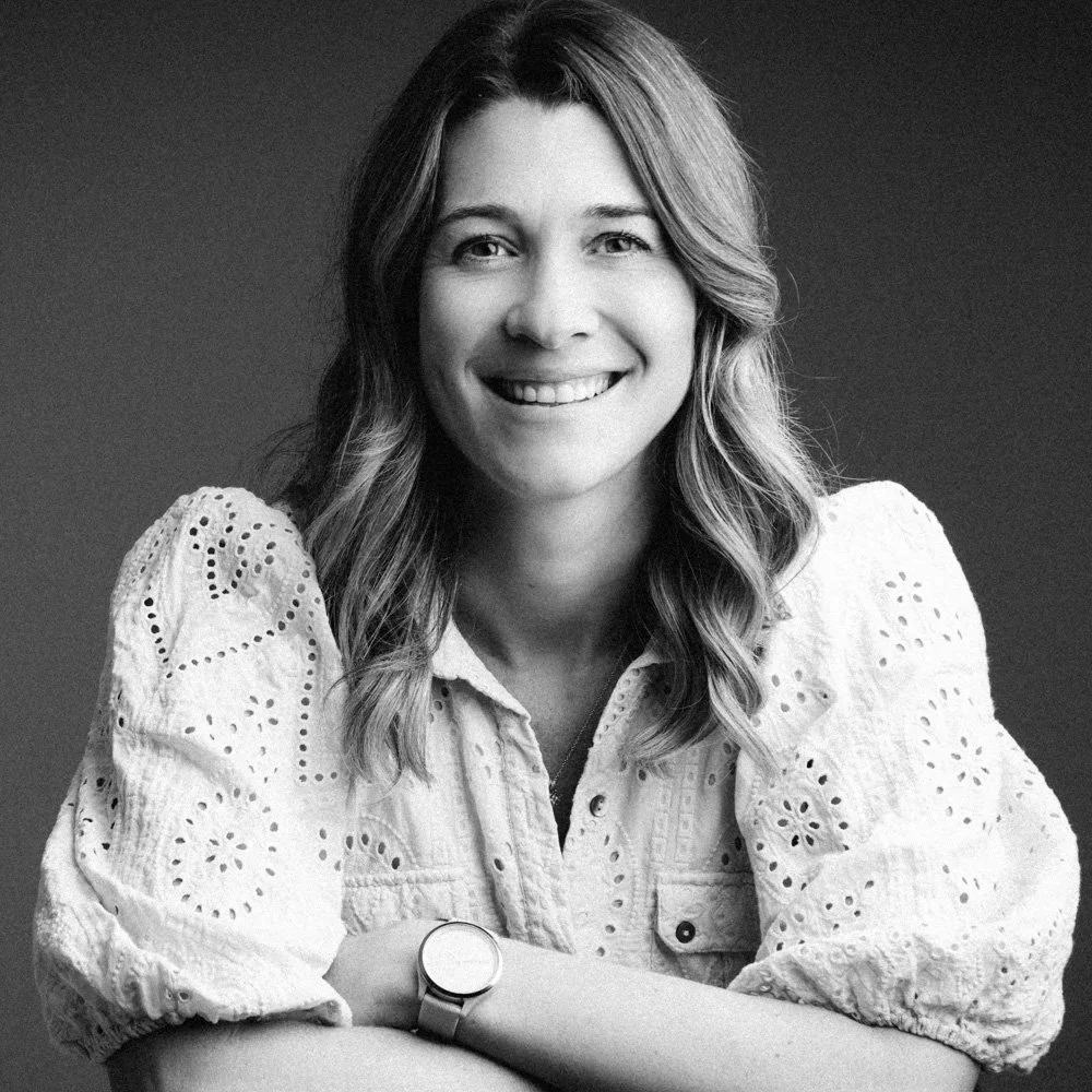 Black and white portrait of a smiling woman with shoulder-length wavy hair, wearing a light-colored embroidered blouse and a watch on her left wrist, against a plain dark background.