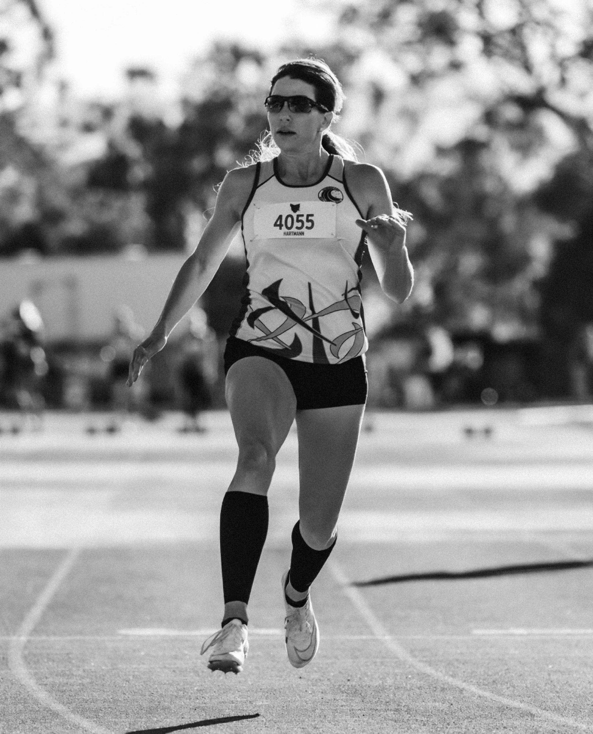 A woman running in a track race, wearing sunglasses, a tank top, shorts, and compression socks with a race bib numbered 4055.