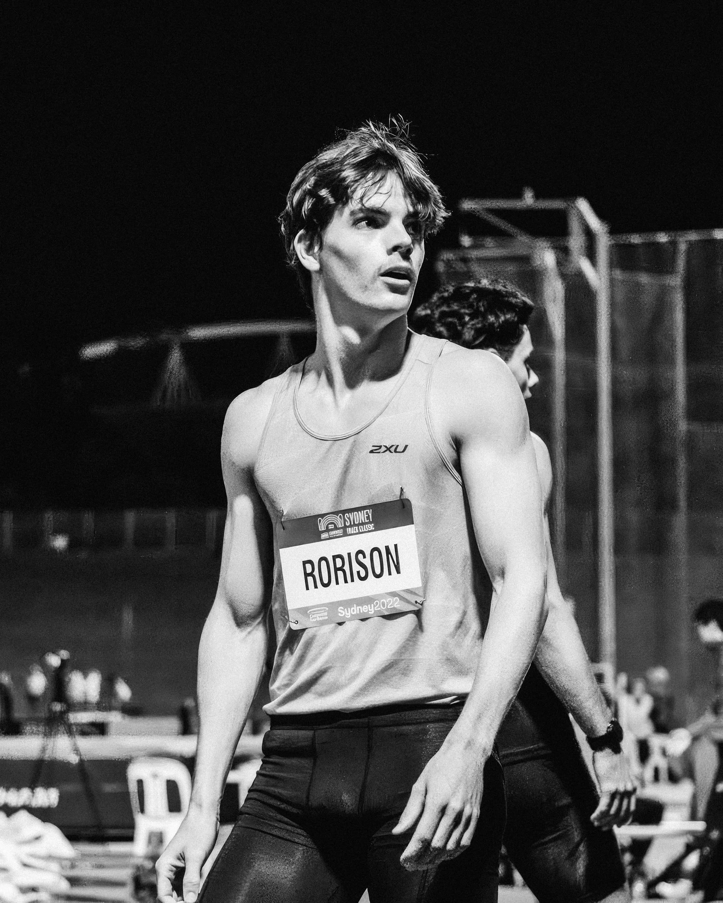 Black and white photo of a male athlete wearing a tank top with a bib number 'RORISON' at a nighttime track event. The athlete has short, tousled hair and a focused expression.