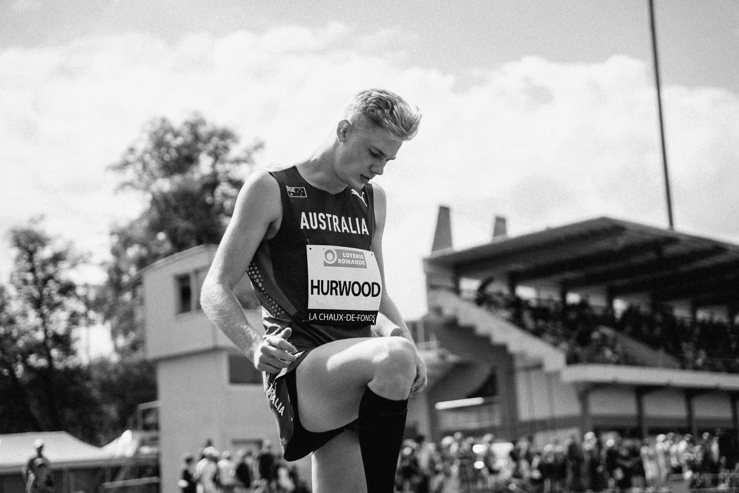A male athlete in a black Australia jersey stretching or warming up on a track at a sports stadium, with spectators and stadium structures in the background.