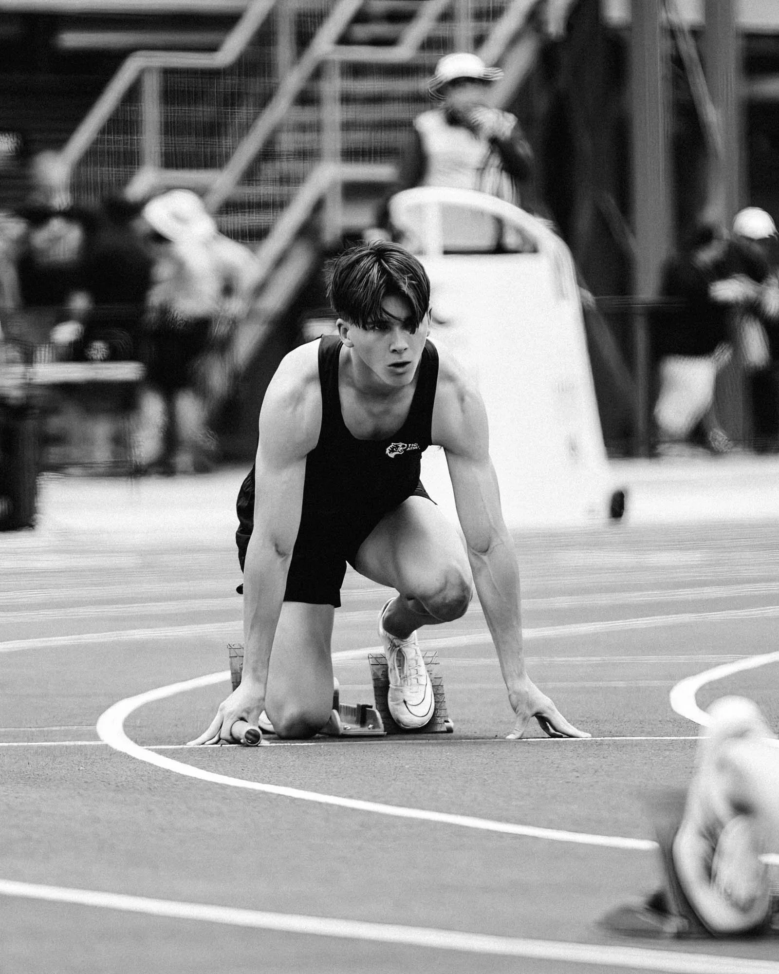 A young male athlete in starting position on a track, preparing for a race, with people and stadium structures in the background.