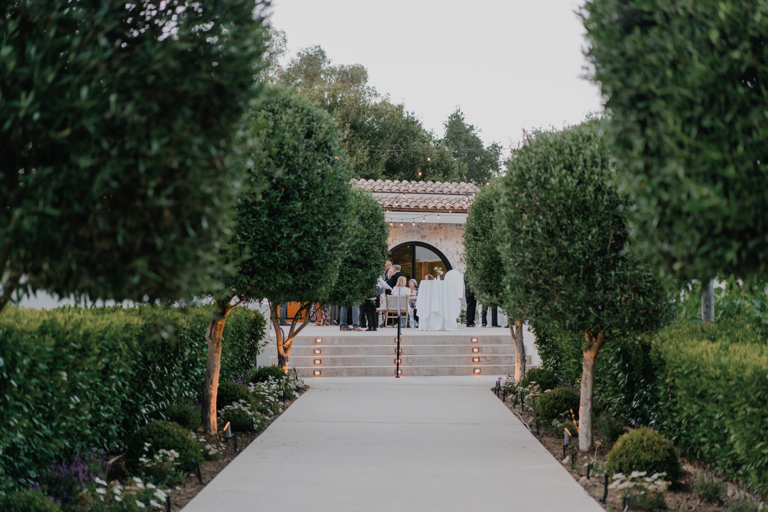 A garden pathway leading to a gathering of people at an outdoor event, with neatly trimmed trees lining the walkway and string lights hanging overhead near a building with a tiled roof.