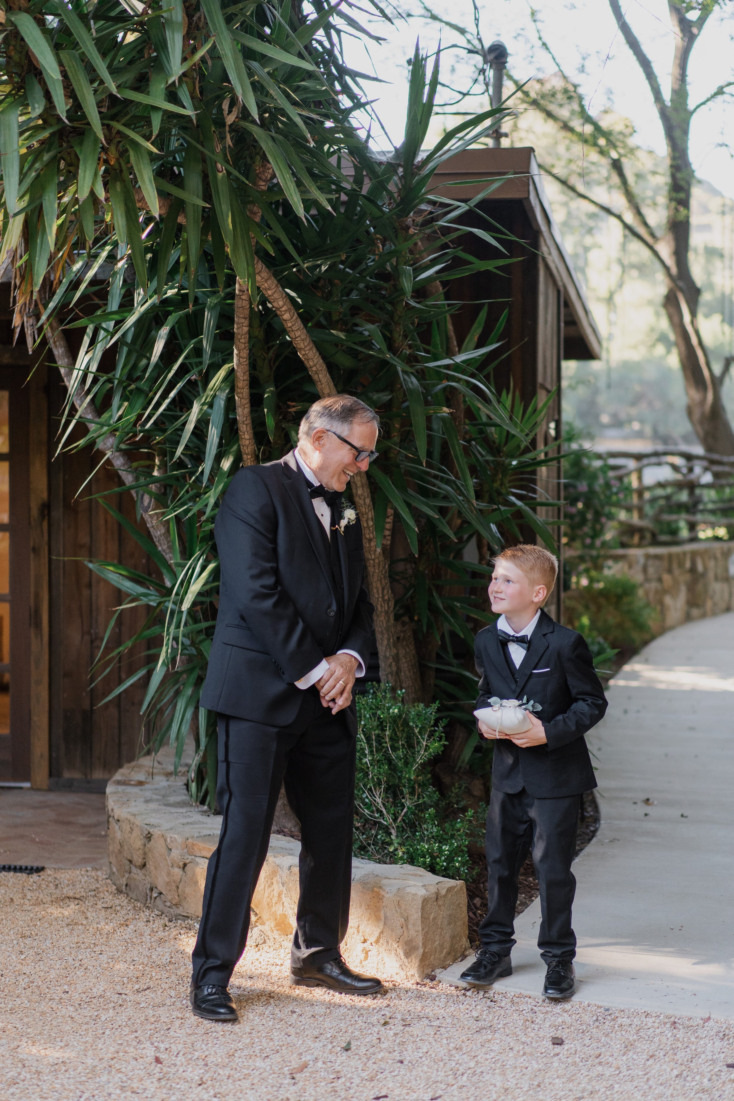 A man and a boy dressed in tuxedos smiling and talking outdoors near a large plant and a wooden building, with sunlight filtering through trees.