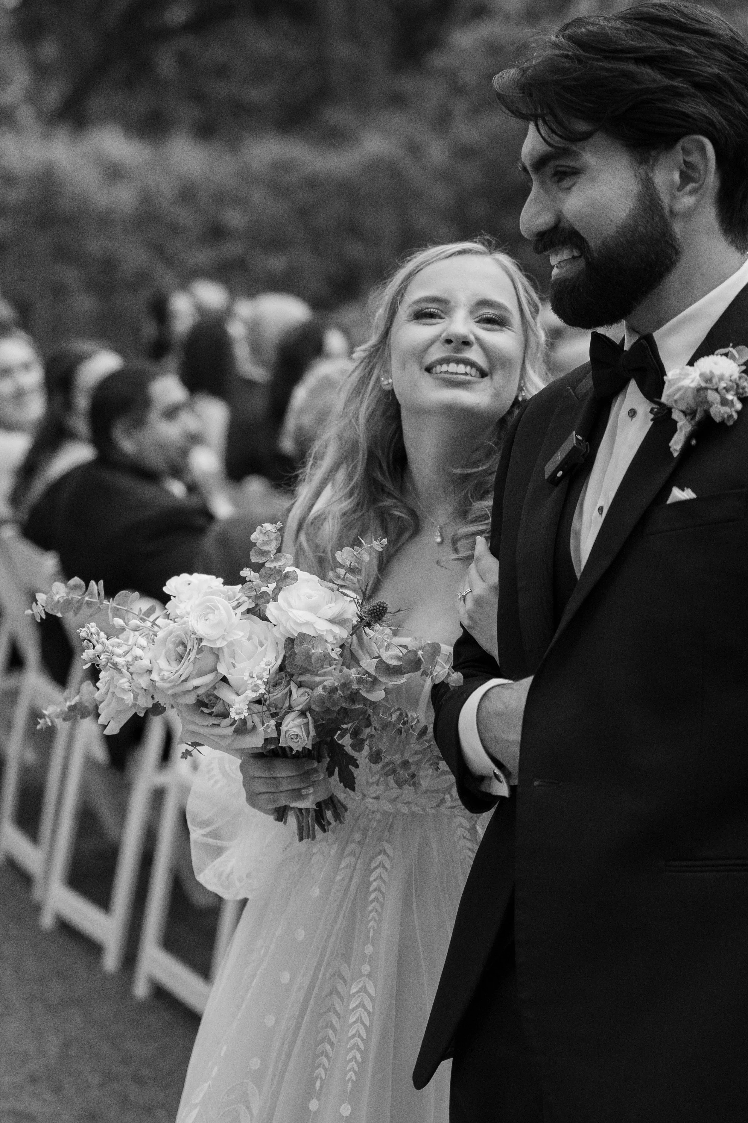 A bride in a wedding dress holding a bouquet of flowers smiles happily at her groom during a wedding ceremony outdoors, with guests seated in the background.