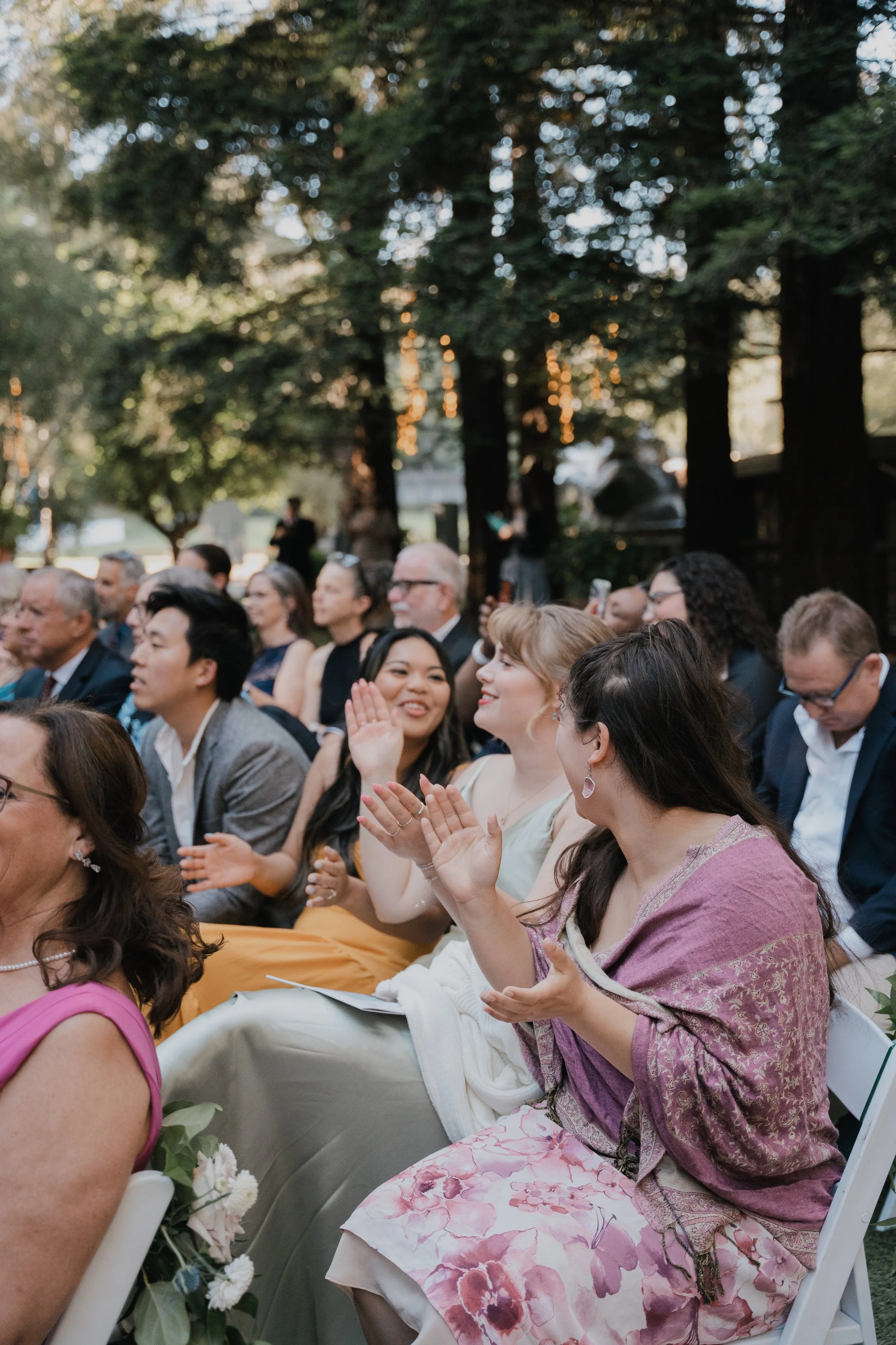 People seated outdoors attending an event, engaged and clapping, with trees in the background.