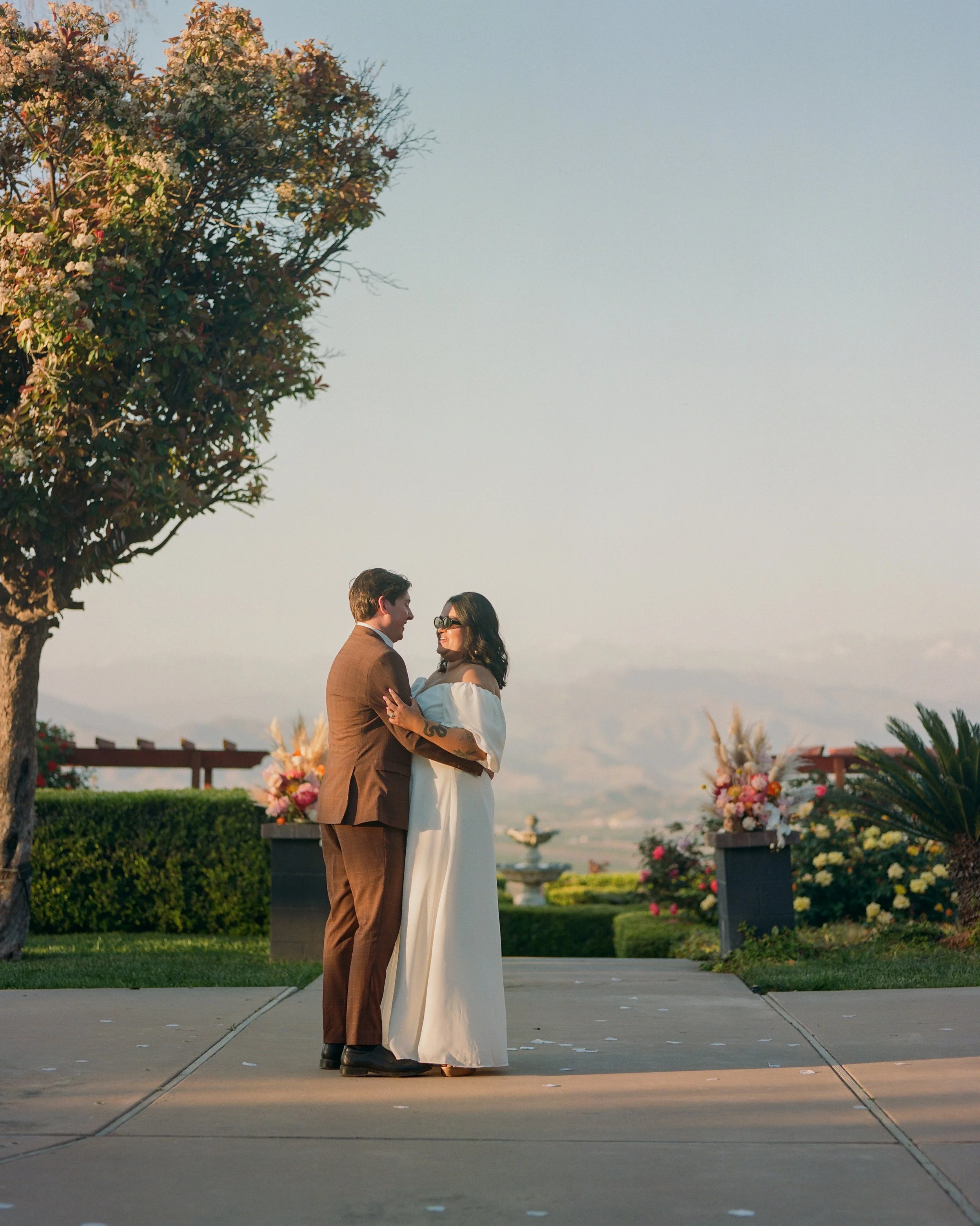 A couple on their wedding day, holding each other and smiling, standing outdoors on a paved path with floral arrangements, greenery, and a sunset sky in the background.