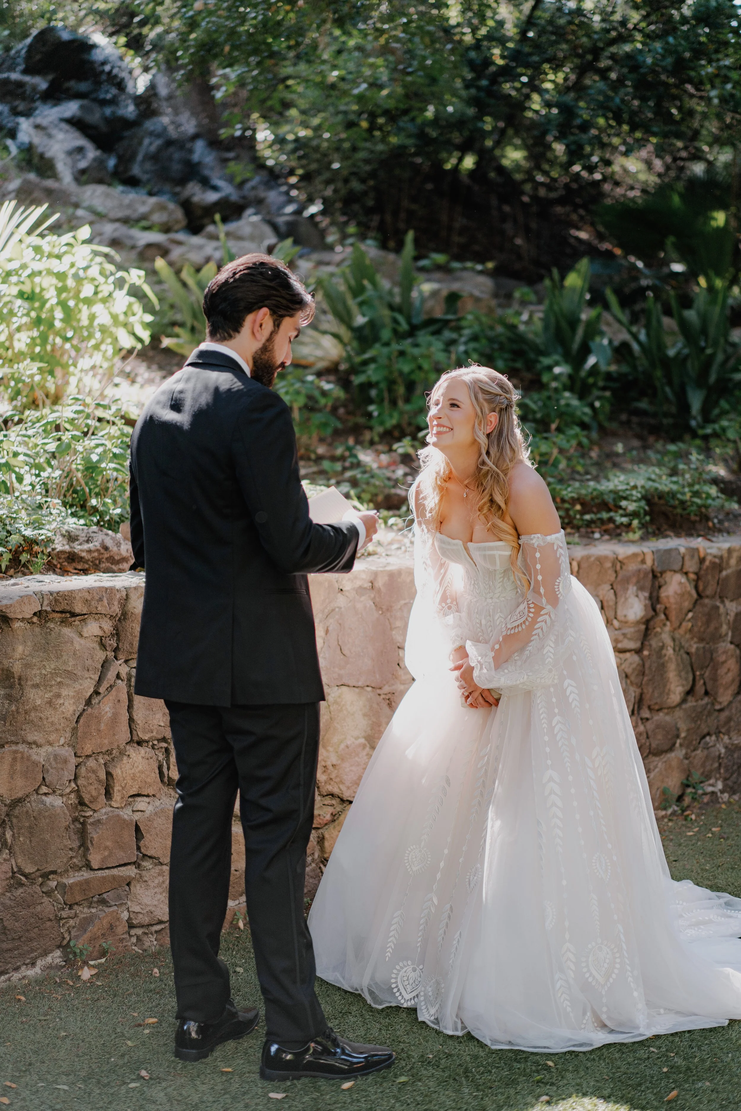 A bride and groom exchange vows outdoors, with the bride smiling and holding hands, in a garden setting.