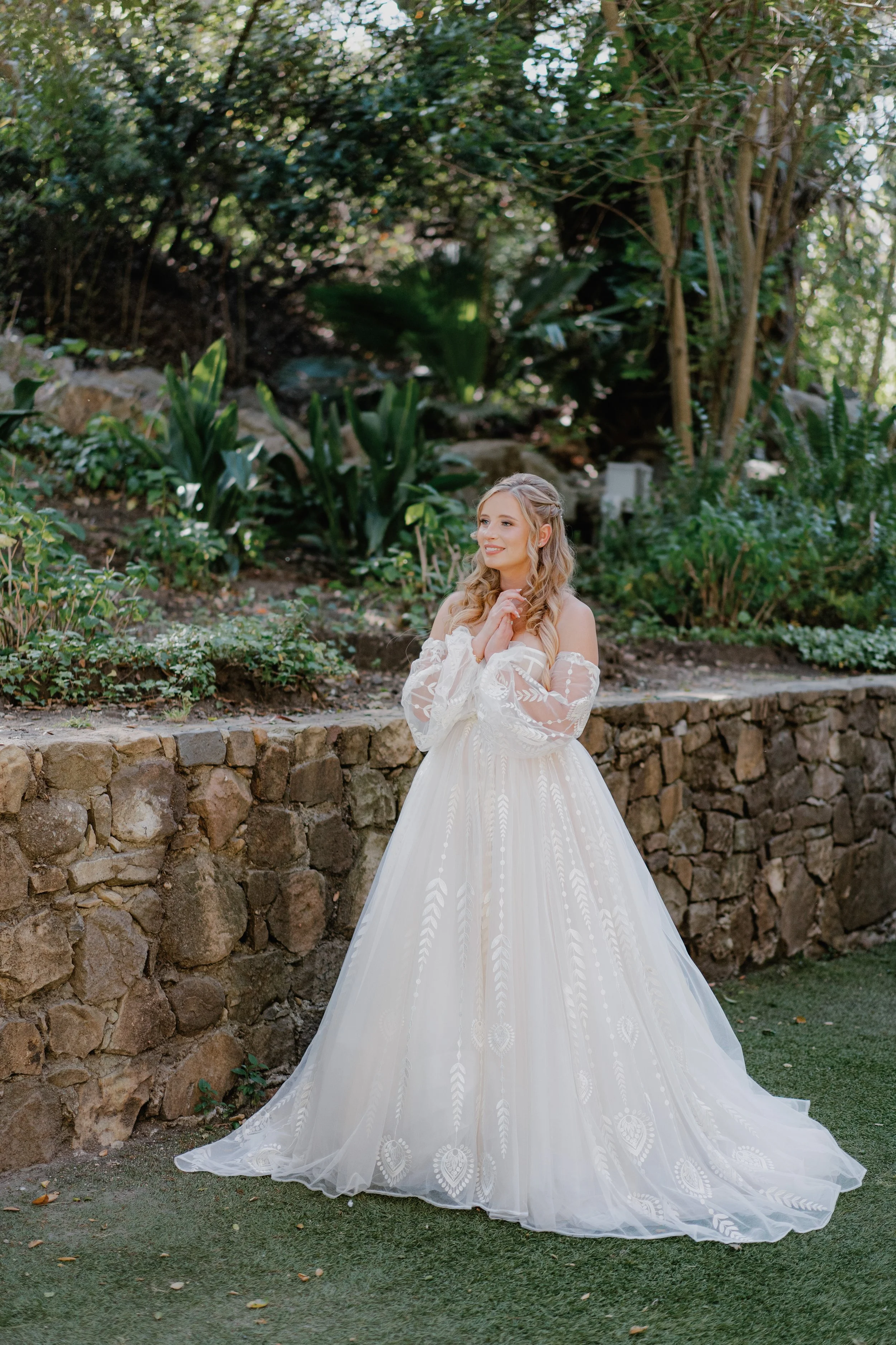 A woman in a white wedding dress standing outdoors on grass near a stone wall, surrounded by greenery and plants, smiling and holding her hands near her chin.