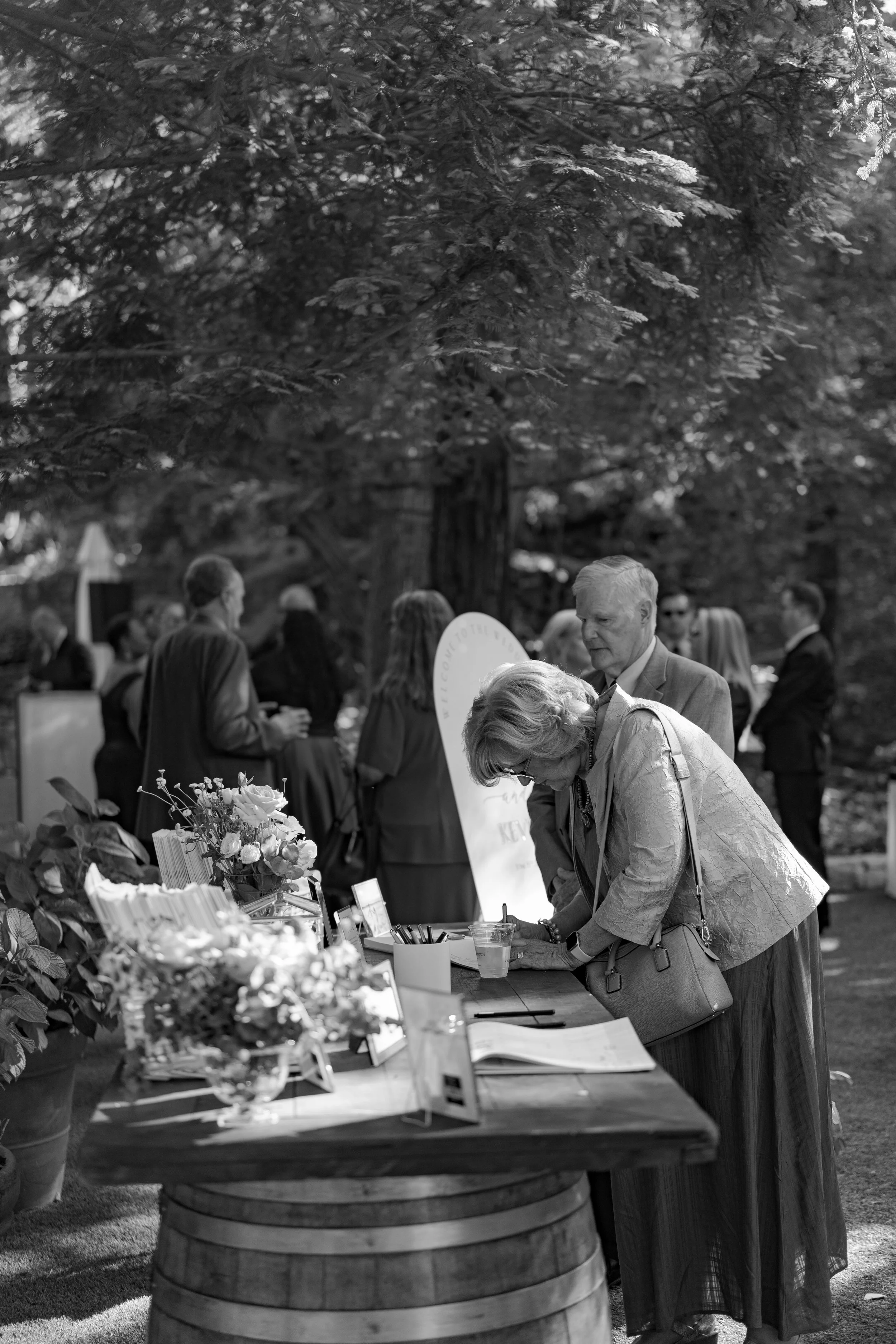 A group of people at an outdoor event, with some gathered around a table with flowers and papers, under a large leafy tree.