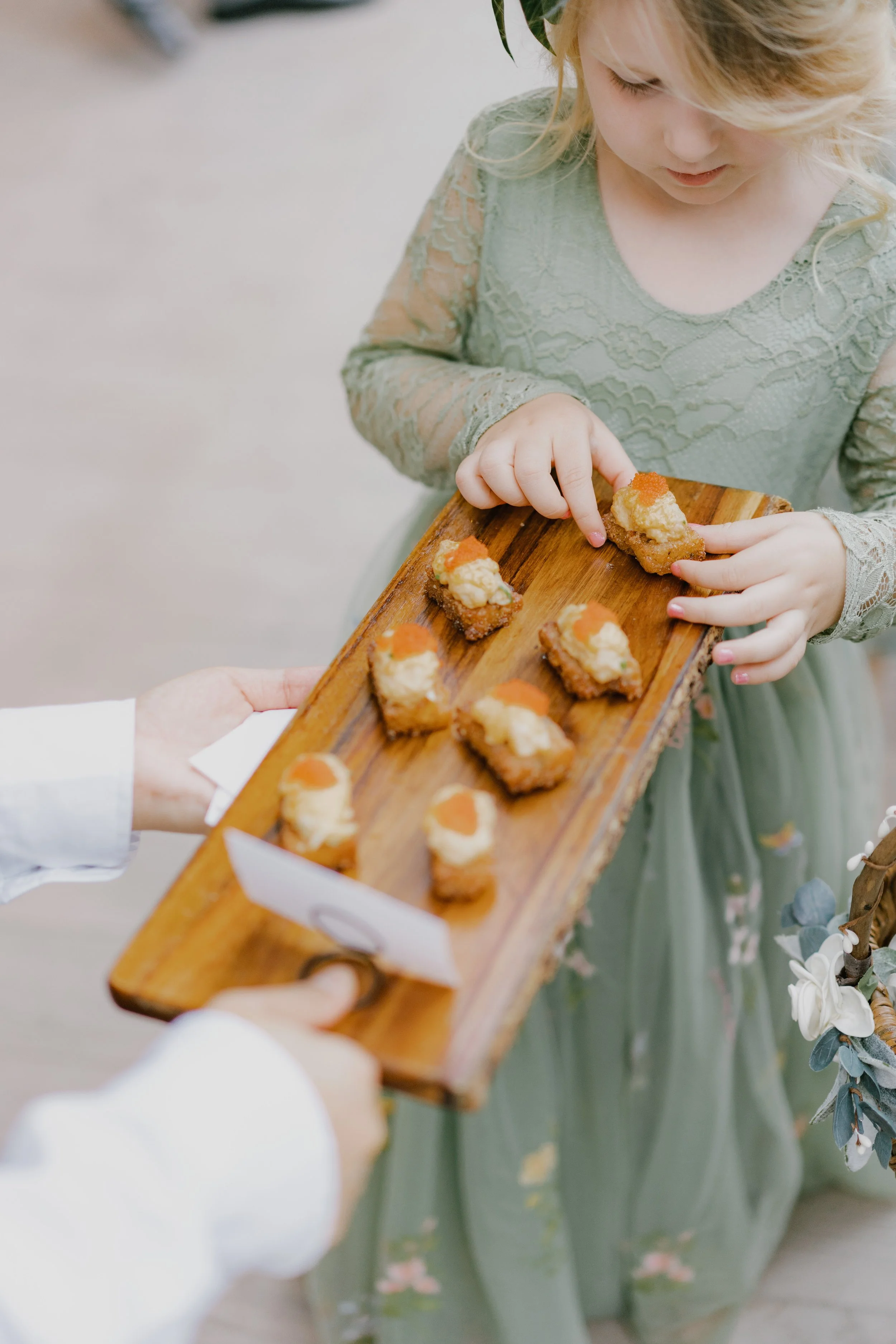 A young girl in a lace green dress receiving a wooden tray of small, topped appetizers from an adult.