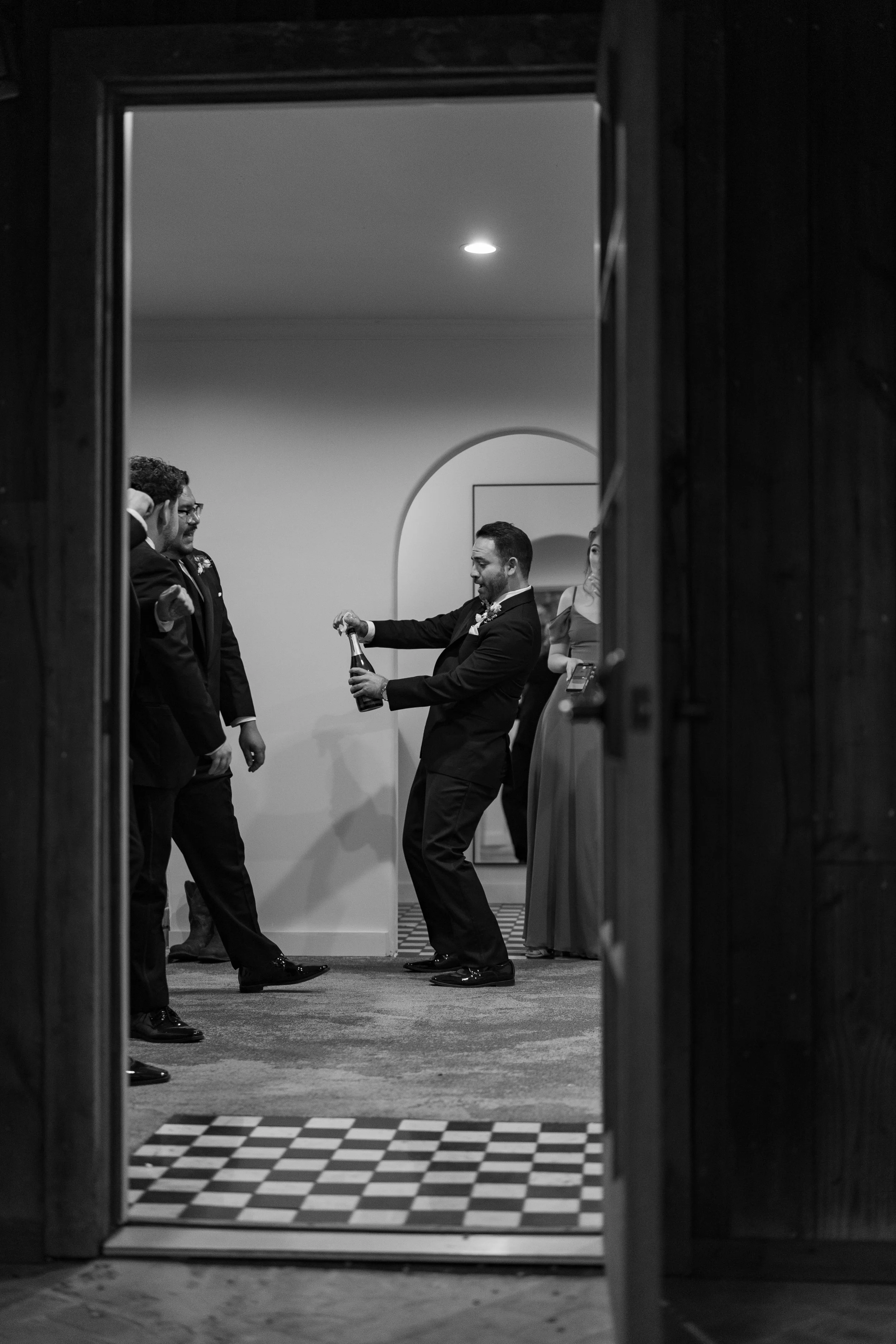 People at a celebration, with one person opening a bottle of champagne and others watching, seen through a doorway in black and white.
