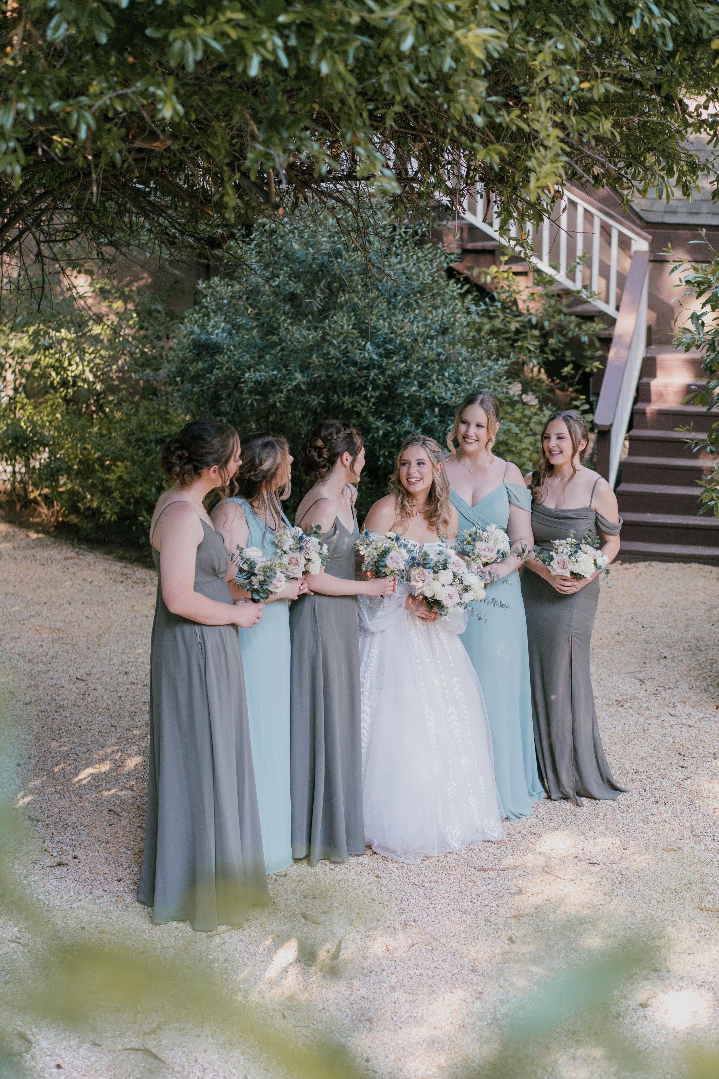 Bride and five bridesmaids standing outdoors, smiling and holding bouquets of flowers, surrounded by greenery.
