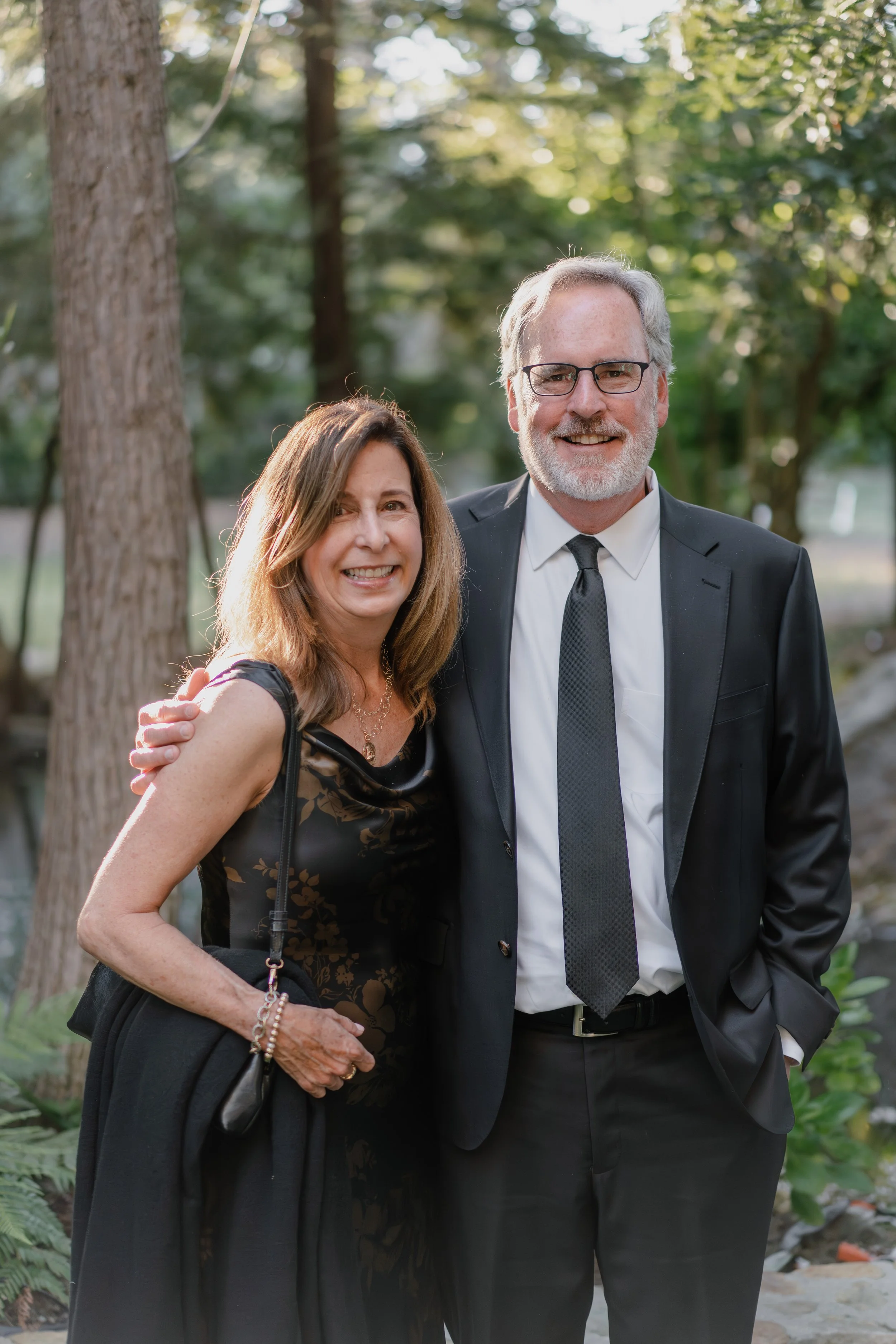 A middle-aged man and woman smiling, standing outdoors in formal attire, with trees and greenery in the background.