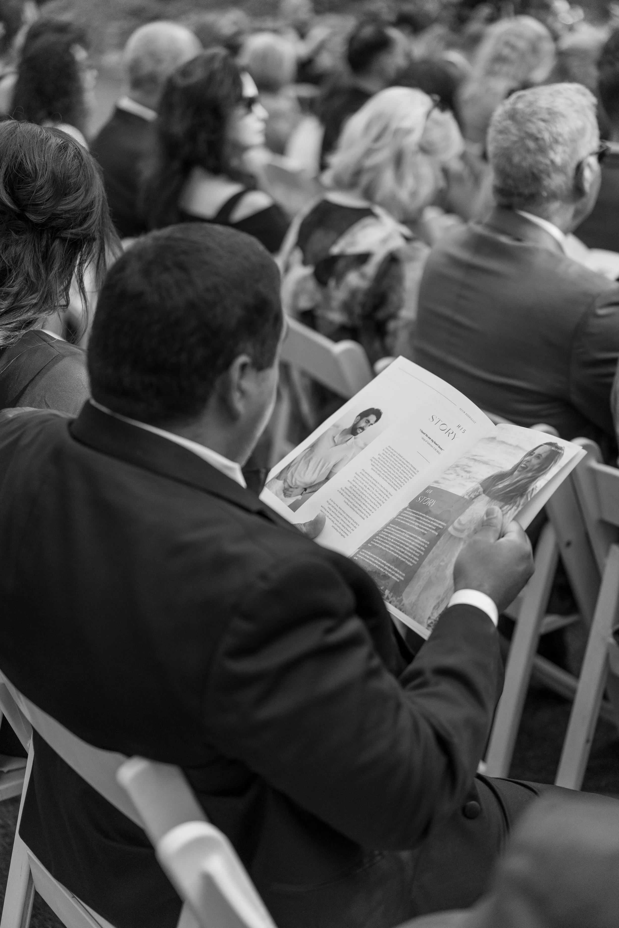 A black-and-white photo of a man in a suit sitting in a chair, reading a magazine during a formal outdoor event attended by other seated people.