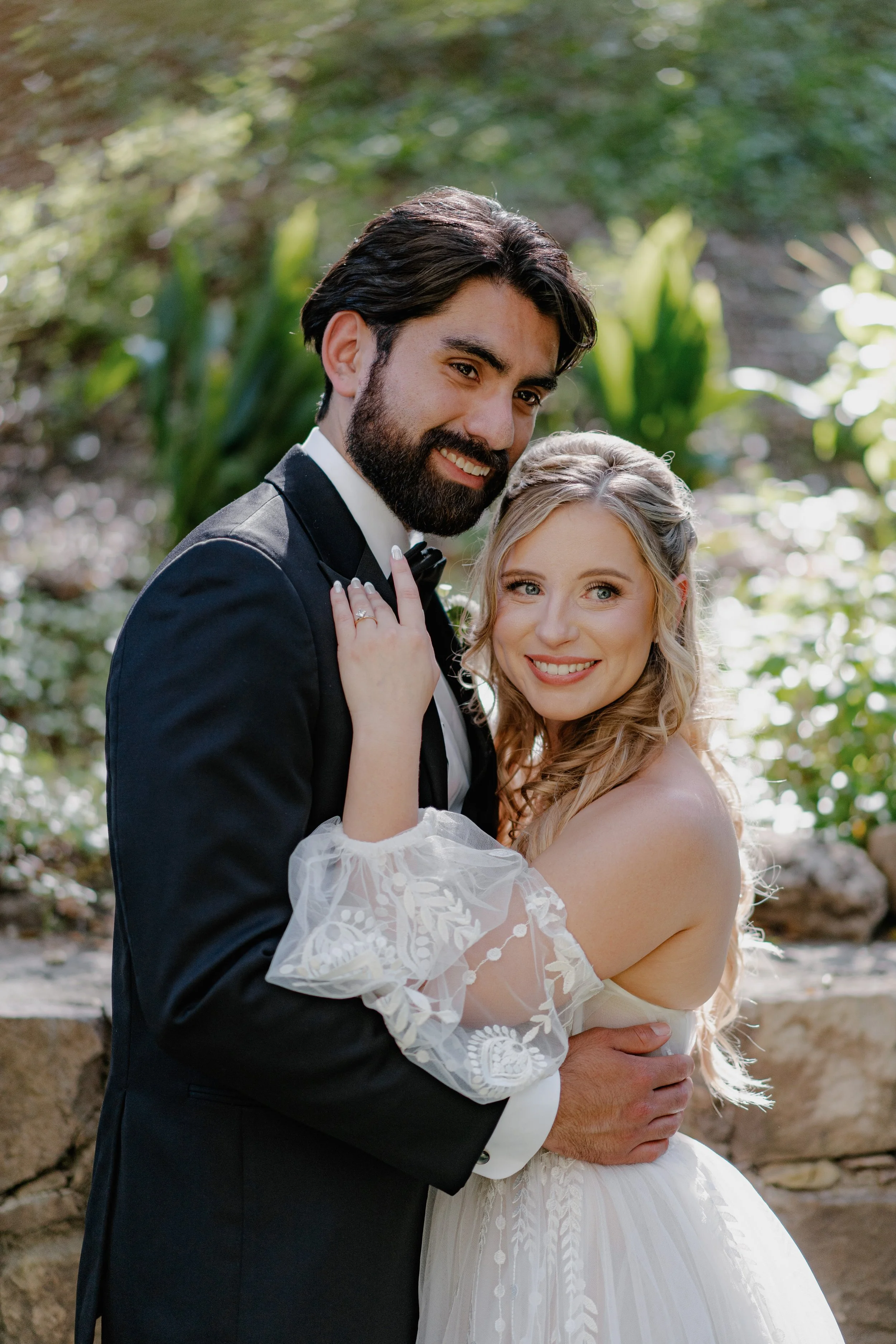 A bride and groom embracing outdoors, the bride in a white dress with lace sleeves, and the groom in a black tuxedo with a bow tie, both smiling at the camera.