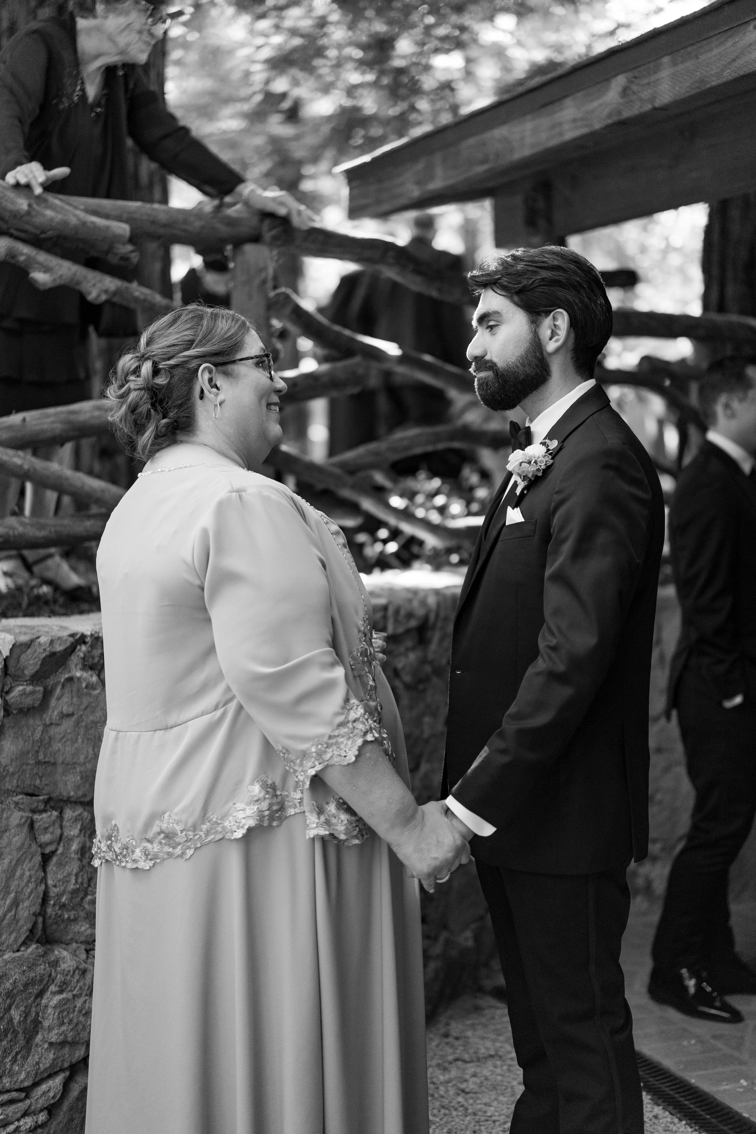 A black and white photo of a wedding ceremony showing a woman and a man holding hands and looking at each other, with the woman smiling, outdoors with wooden and stone structures in the background.