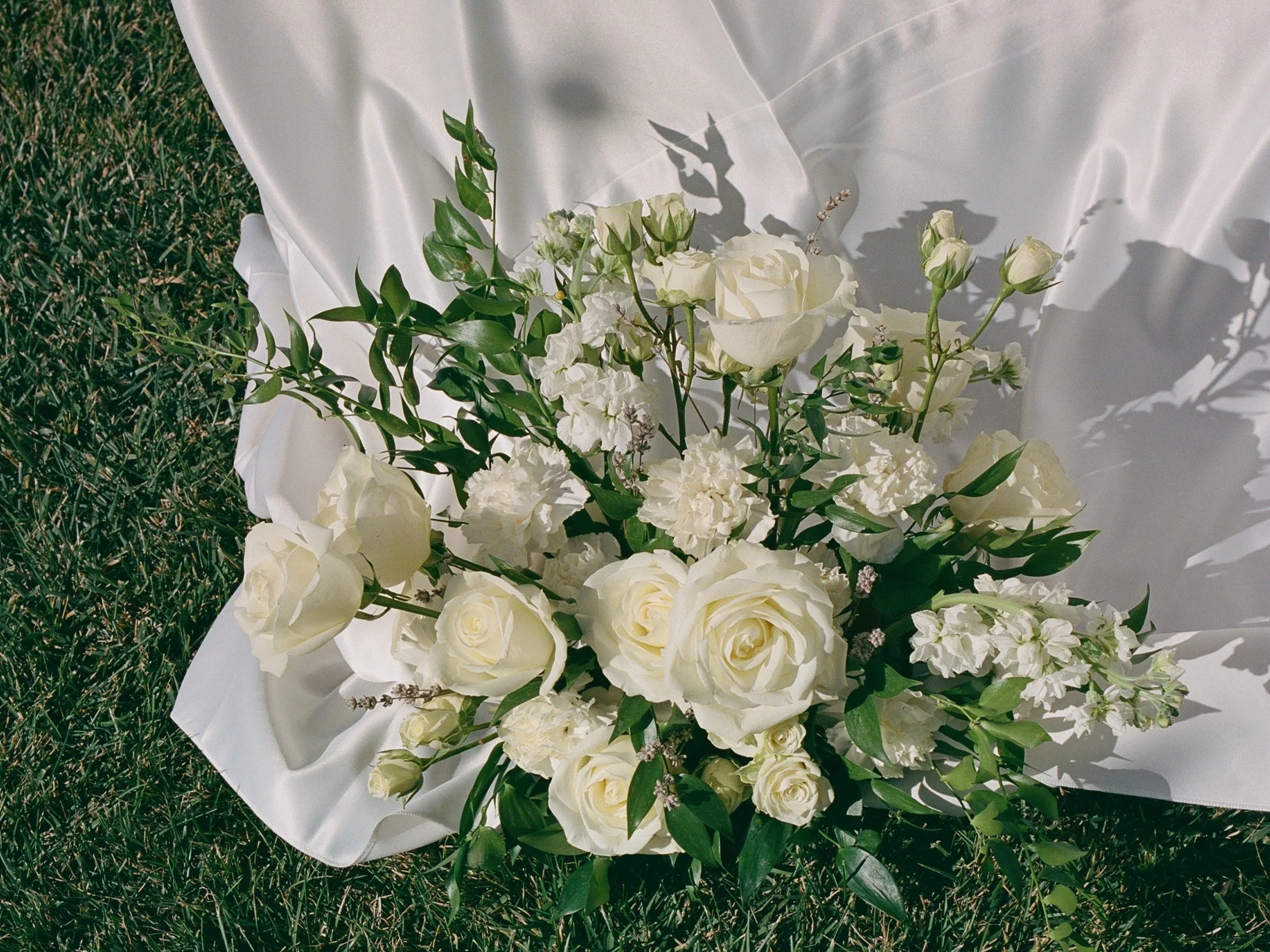 A bouquet of white roses, lisianthus, eucalyptus, and other flowers, arranged on white satin fabric on grass.