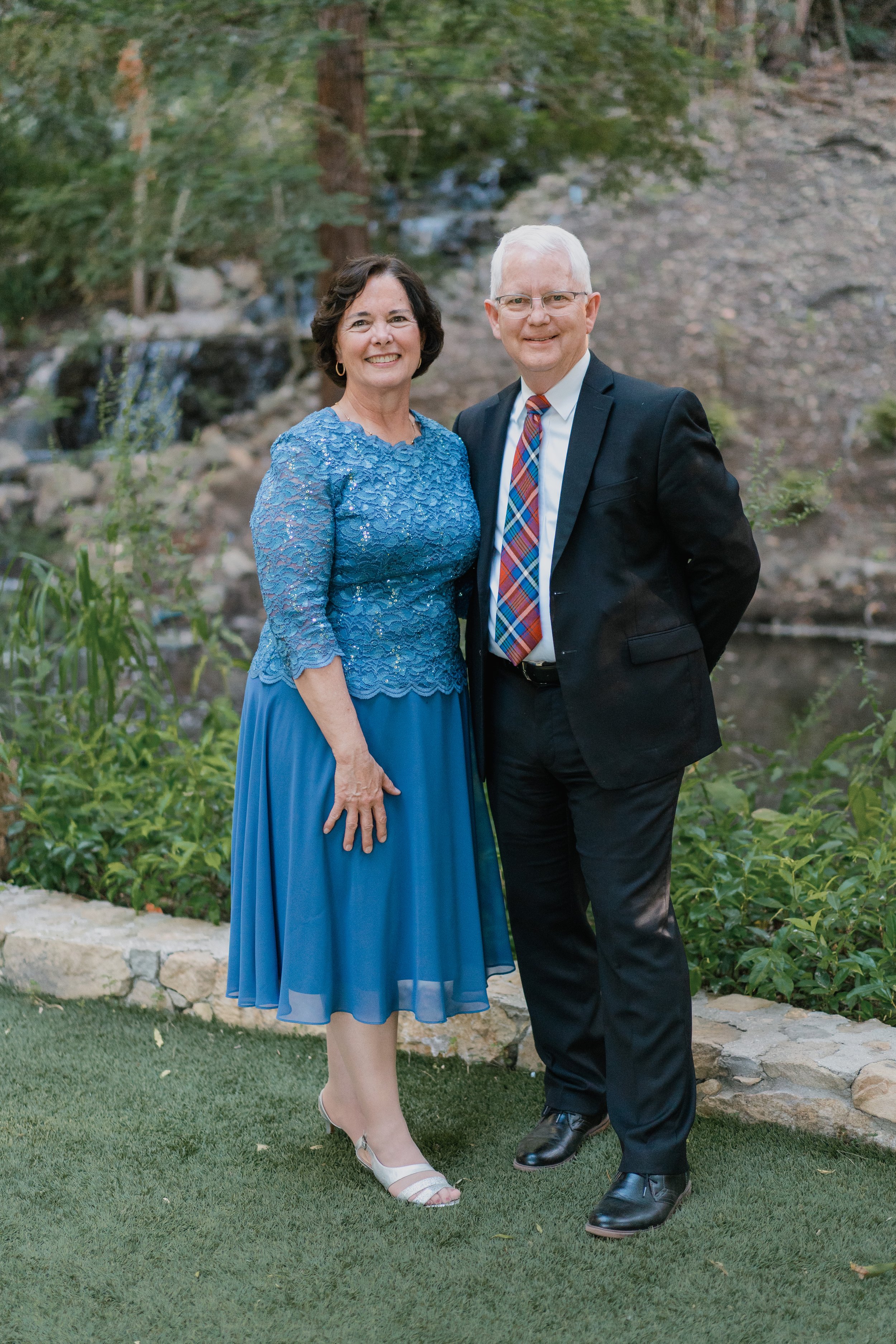 A smiling couple standing outdoors near a small creek, with lush greenery and rocks in the background. The woman is wearing a blue lace dress and heels, and the man is dressed in a black suit with a plaid tie and dress shoes.