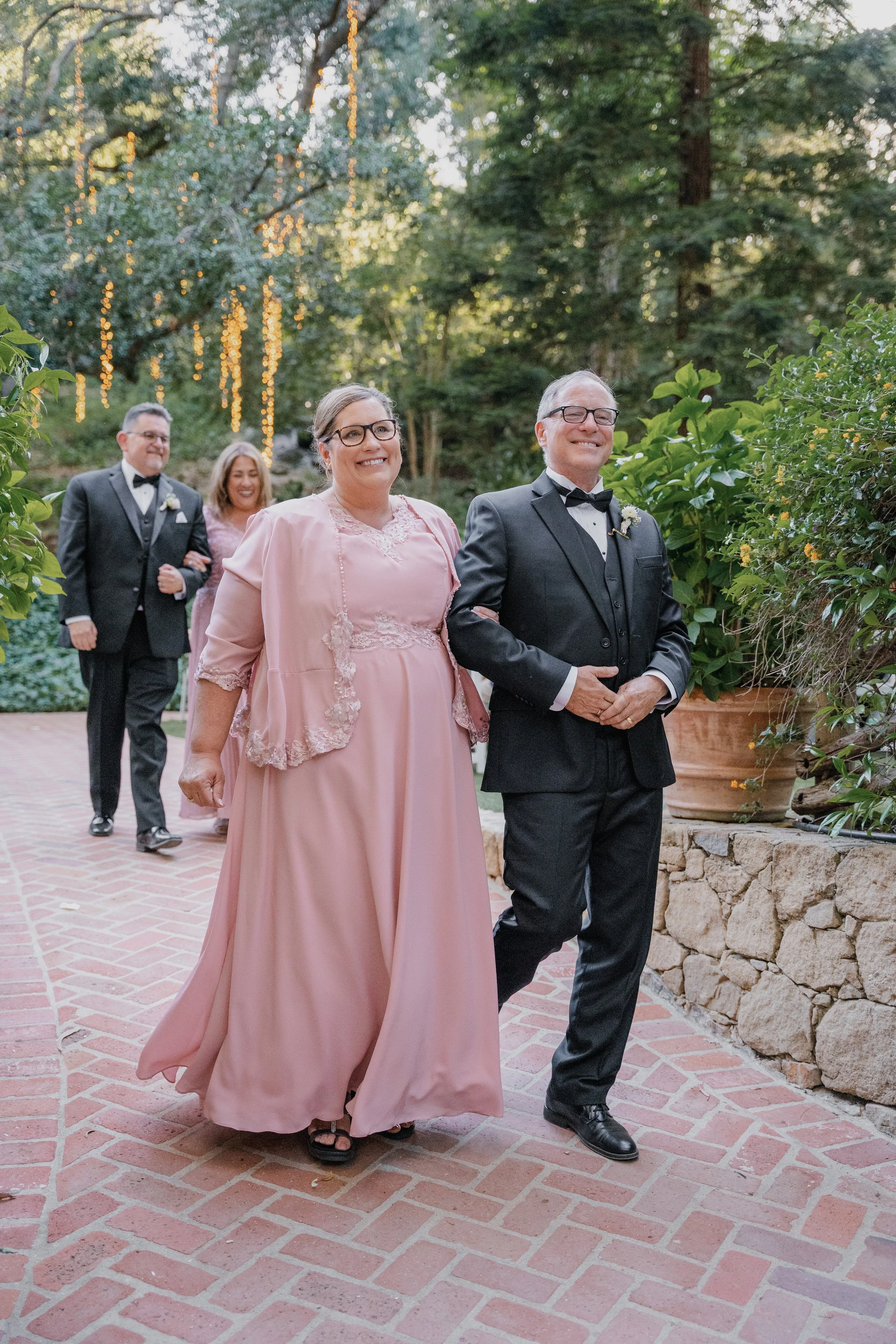 Group of people walking outdoors on a brick path, dressed in formal attire, with string lights and greenery in the background
