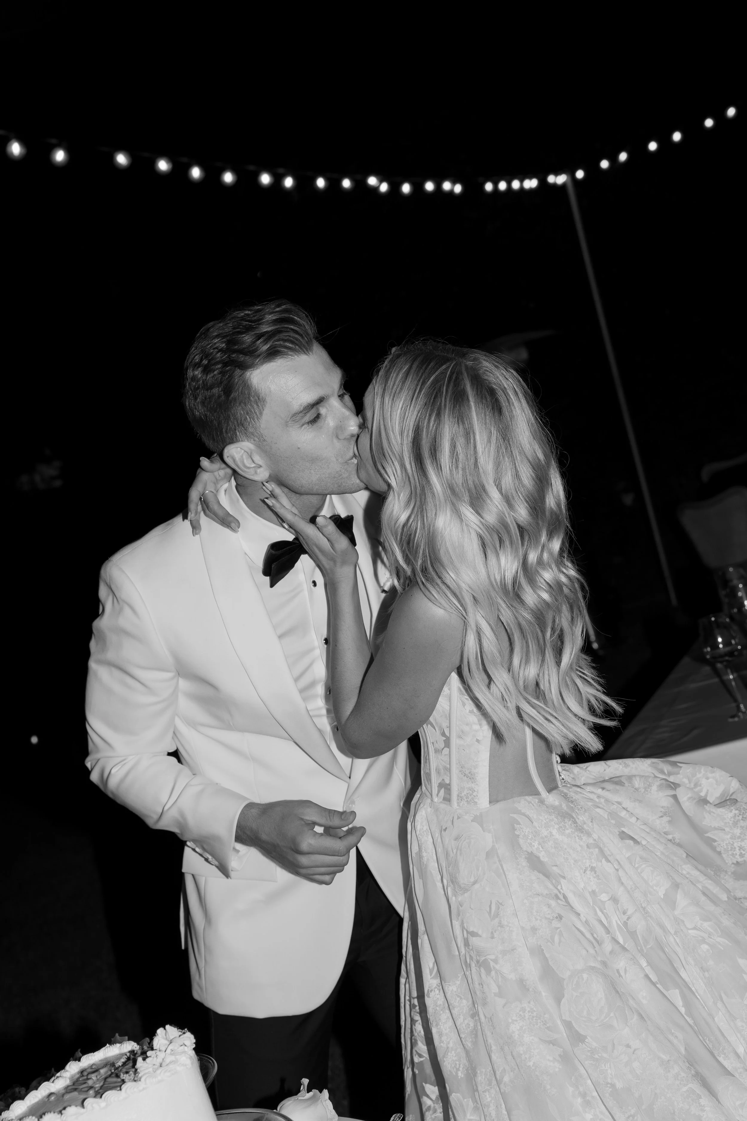 Black and white photo of a bride and groom sharing a kiss at their wedding reception, with string lights in the background.