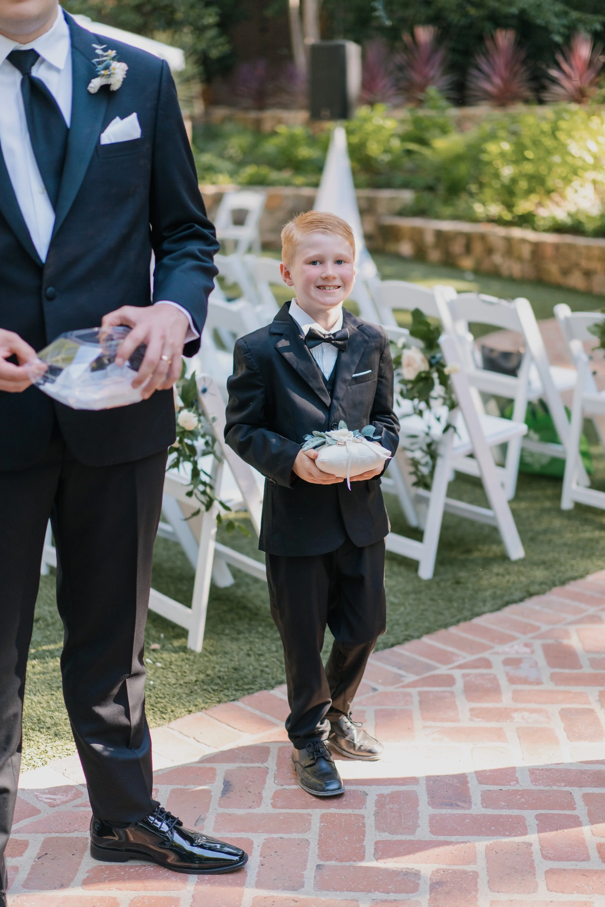 Young boy in a black tuxedo and bow tie holding a small pillow, standing outdoors at a wedding setting, with empty white chairs decorated with flowers behind him.