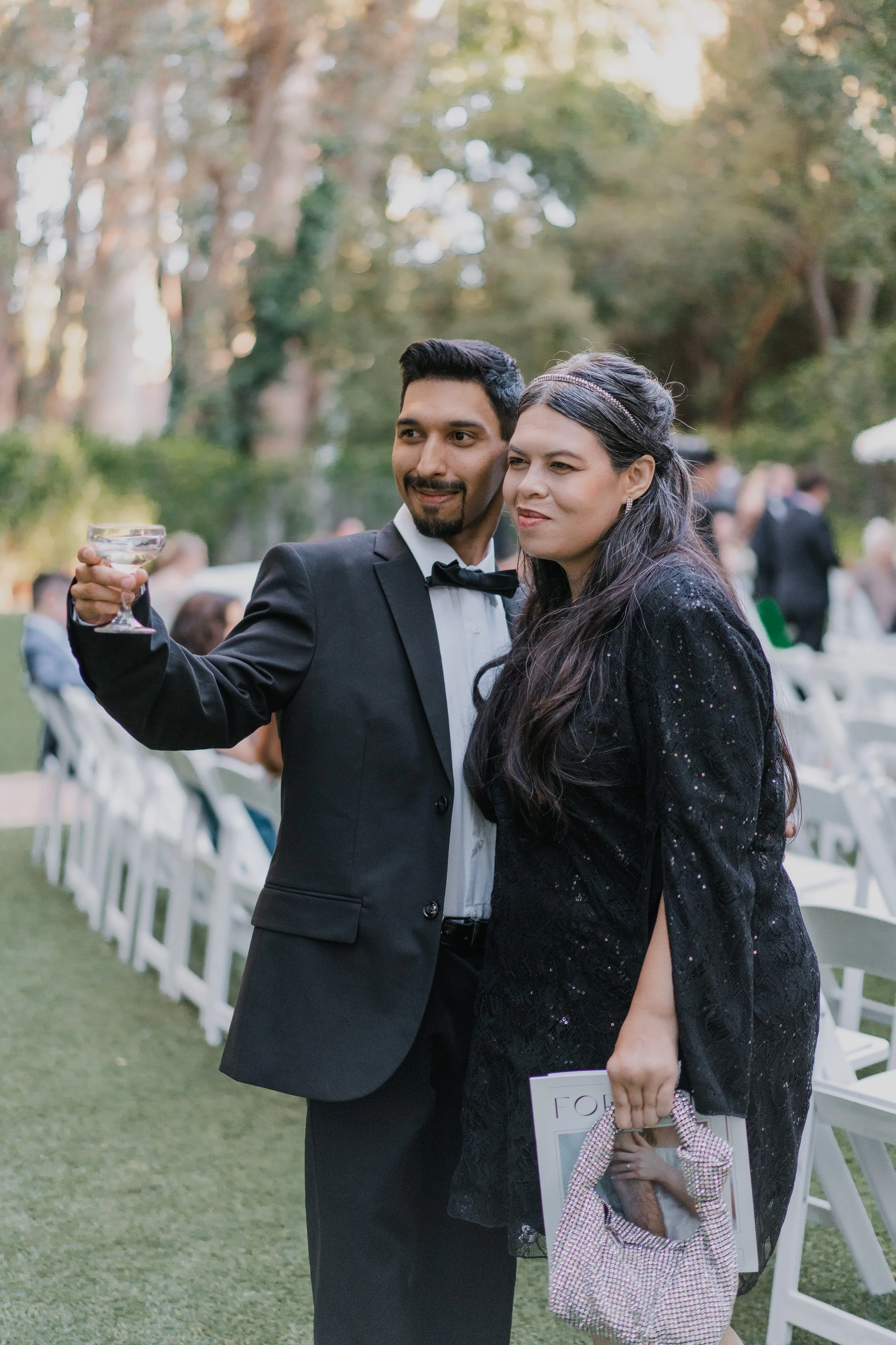 A man in a tuxedo holding a glass, standing next to a woman in a black dress holding a magazine and a small bag, at an outdoor formal event with white chairs and trees in the background.