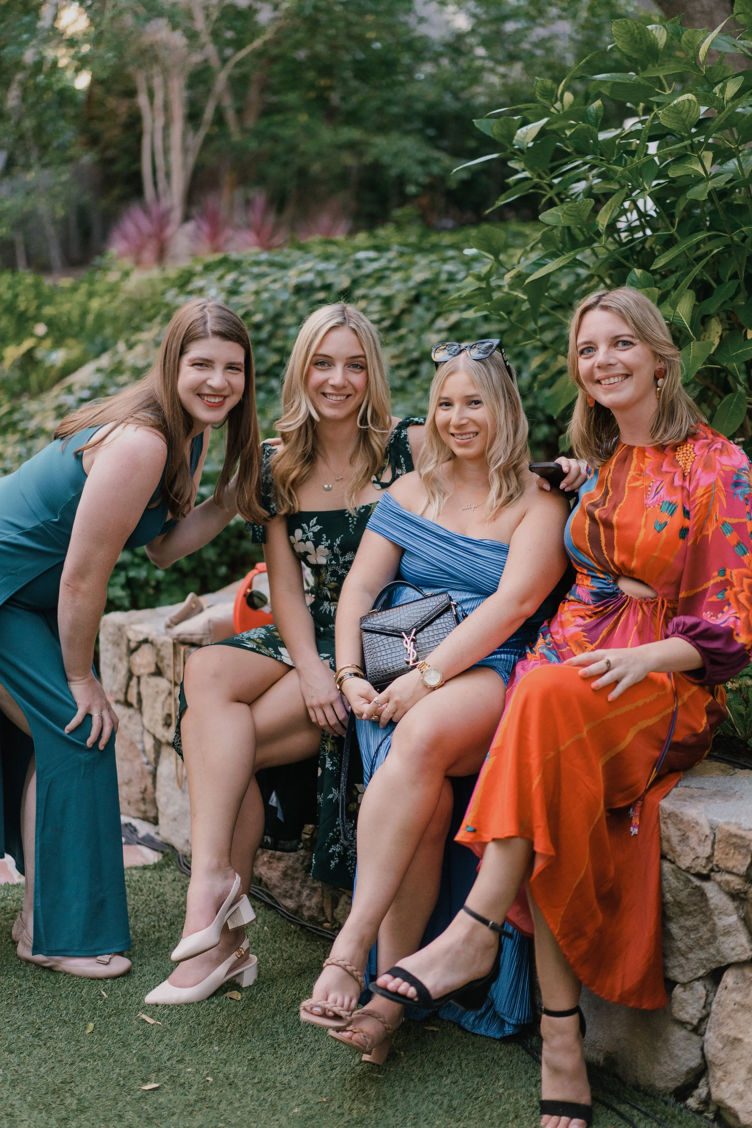 Four women sitting outdoors on a stone wall, smiling at the camera, surrounded by greenery and plants.