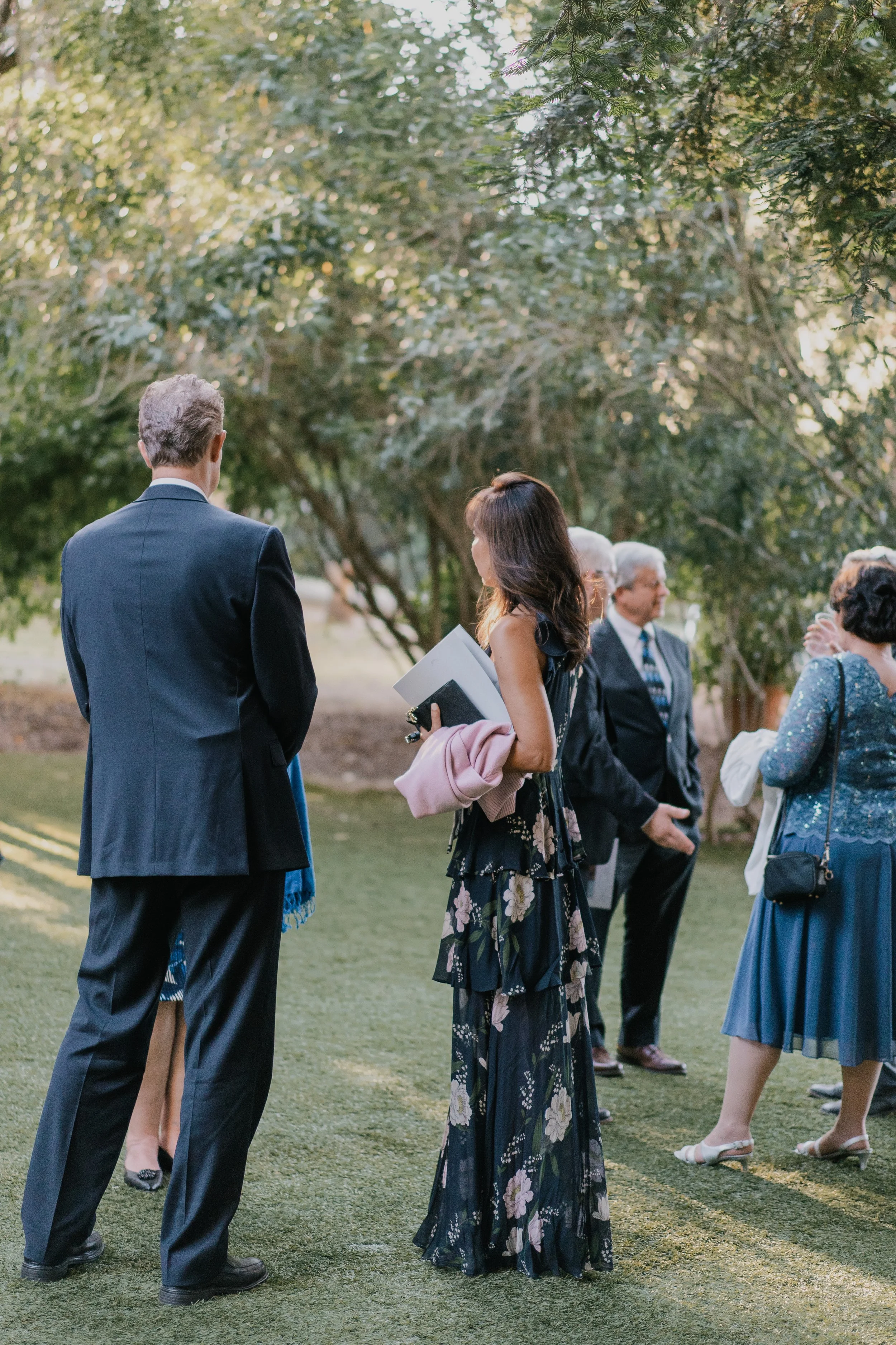 People dressed in formal attire, standing outdoors on grass, engaged in conversation under trees at event or gathering.