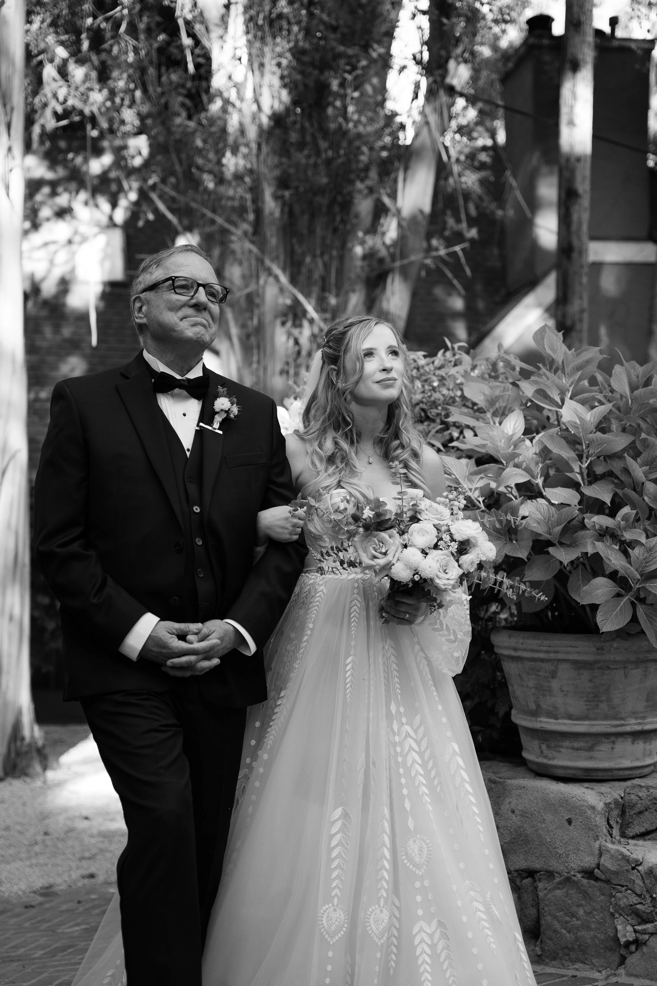 Bride walking down the aisle with her father during a wedding ceremony outdoors, both dressed in formal attire, with the bride holding a bouquet of flowers.