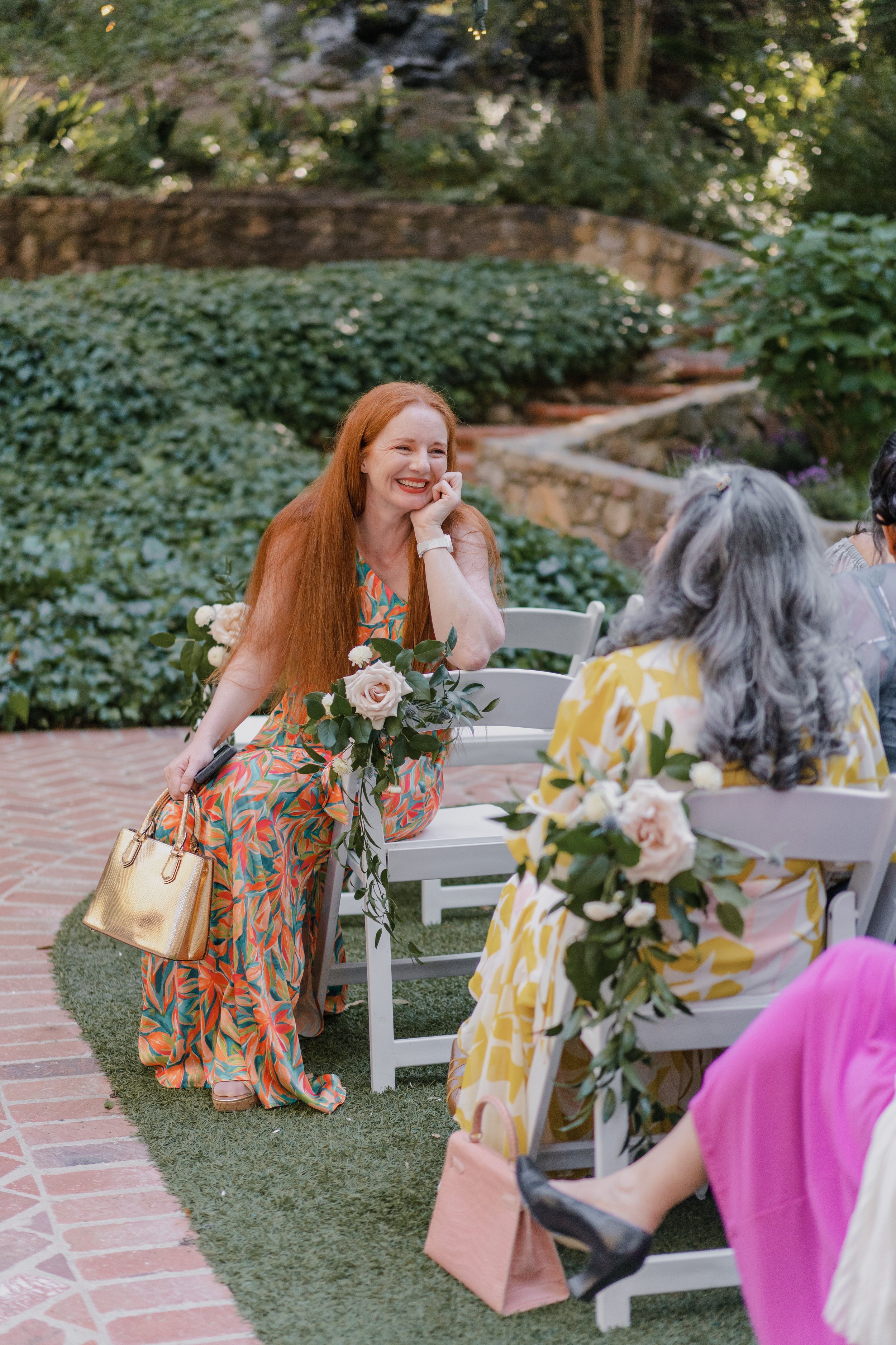 A woman with long red hair wearing a colorful floral dress seated at a table during an outdoor event, smiling and leaning on her hand, with a bouquet of flowers on the table.