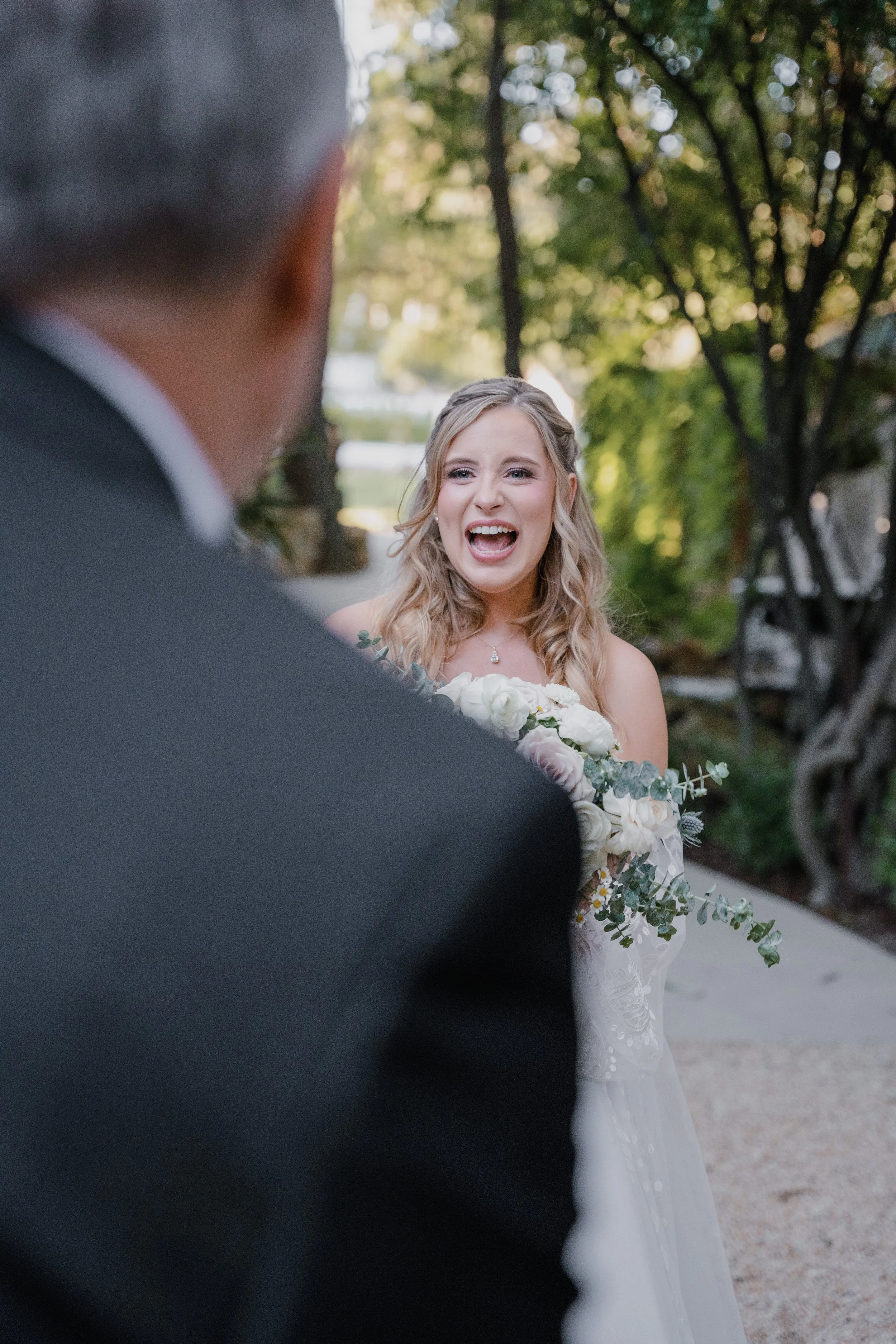 A bride with blonde wavy hair, wearing a white wedding dress, and holding a bouquet of white flowers and greenery, looks emotional and happy as she sees her groom for the first time outdoors. The groom is out of focus in the foreground, wearing a tux